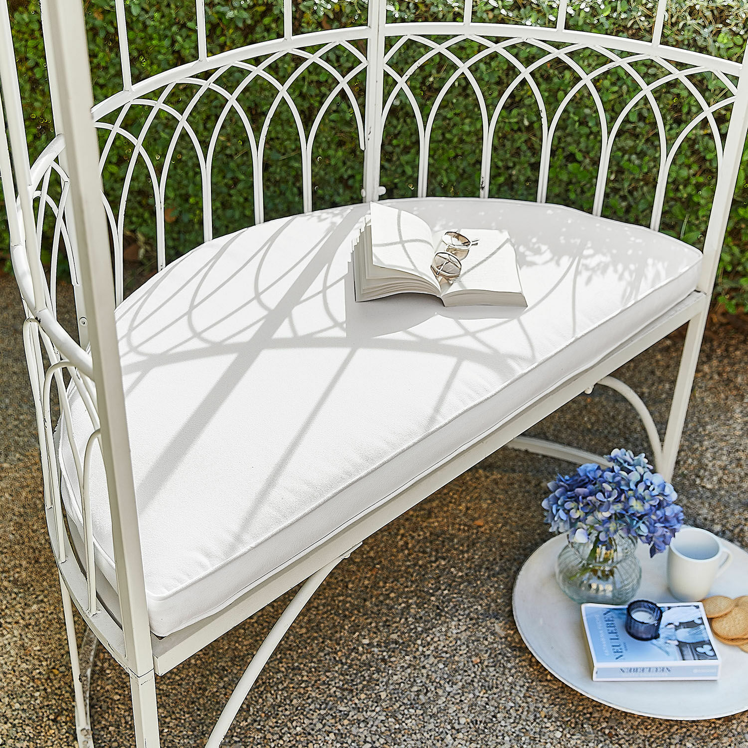Cojín de asiento Drivesto Banco de jardín metálico blanco con cojín blanco, libro abierto, flores azules y taza en terraza