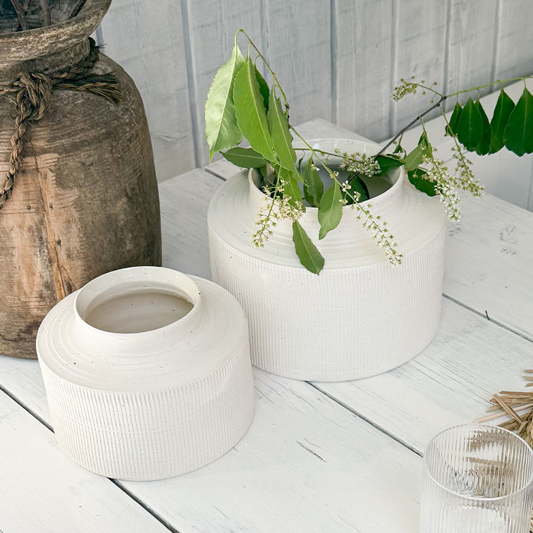 Jarrones de cerámica blanca mate sobre mesa de madera con hojas verdes y flores blancas