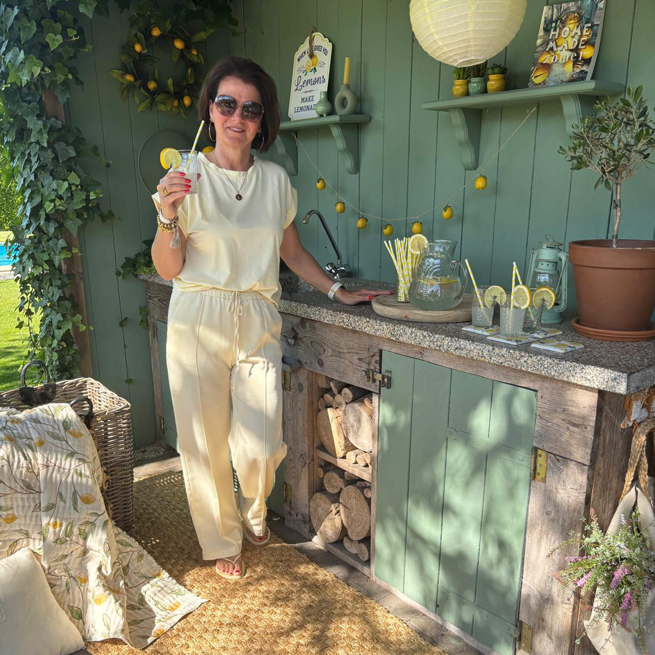 Mujer con conjunto crema junto a cocina exterior verde con limonada y decoración de limones