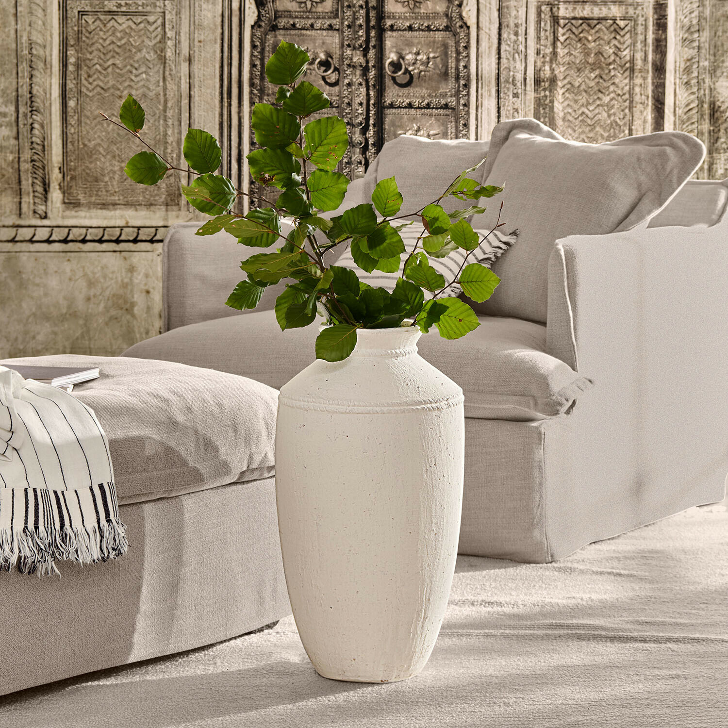 Jarrón de cerámica blanca con hojas verdes frente a un sofá beige y pared decorativa en salón