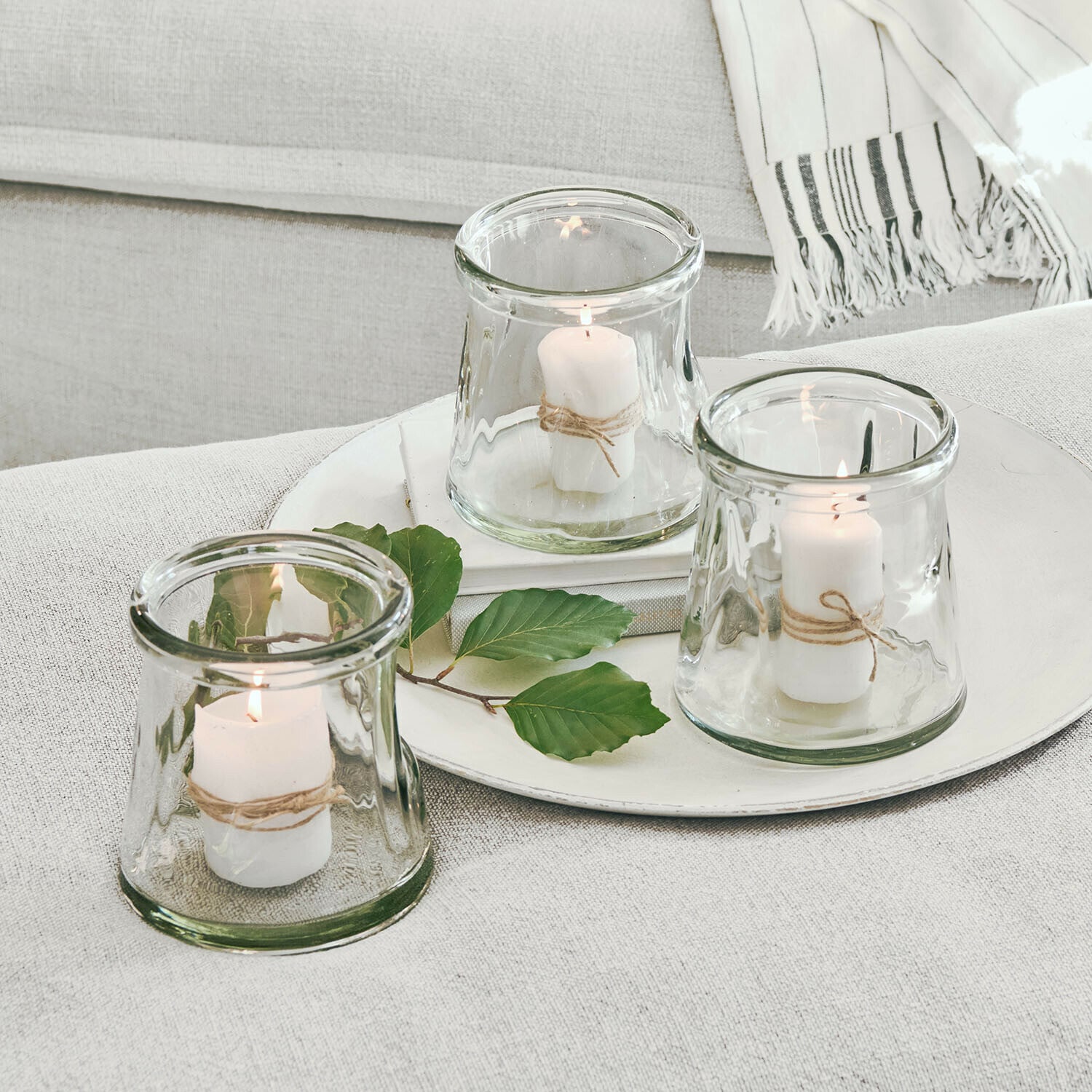 Set de 3 portavelas Gravière Conjunto de tres portavelas de vidrio con velas blancas y cuerda natural sobre fondo claro