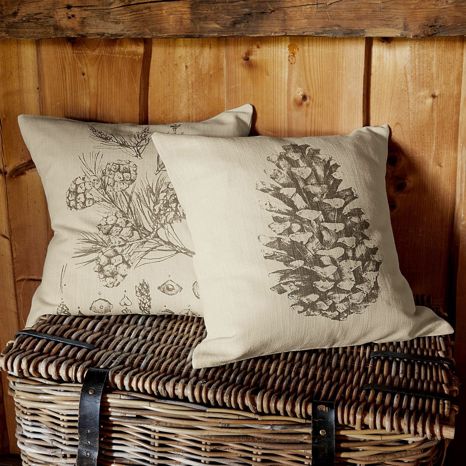 Cojines de lino beige con estampado de piñas y ramas sobre cesta de mimbre, decoración natural