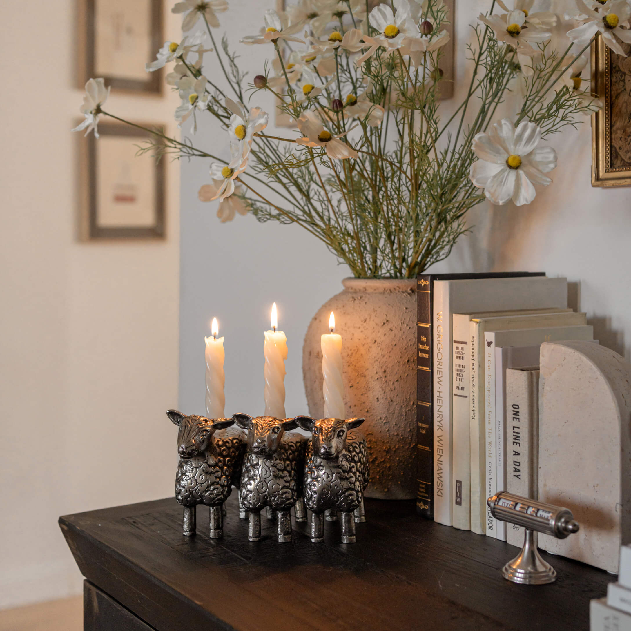 Candelabro metálico con forma de tres ovejas con velas blancas sobre mesa de madera oscura junto a libros