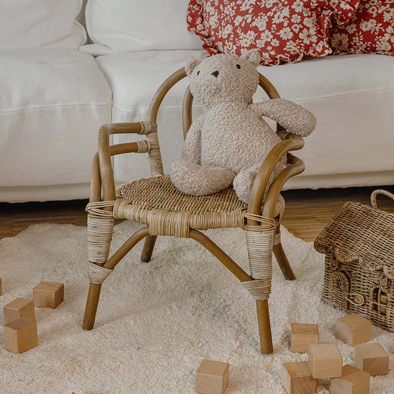 Sillita infantil de ratán con oso de peluche beige sobre alfombra clara y bloques de madera