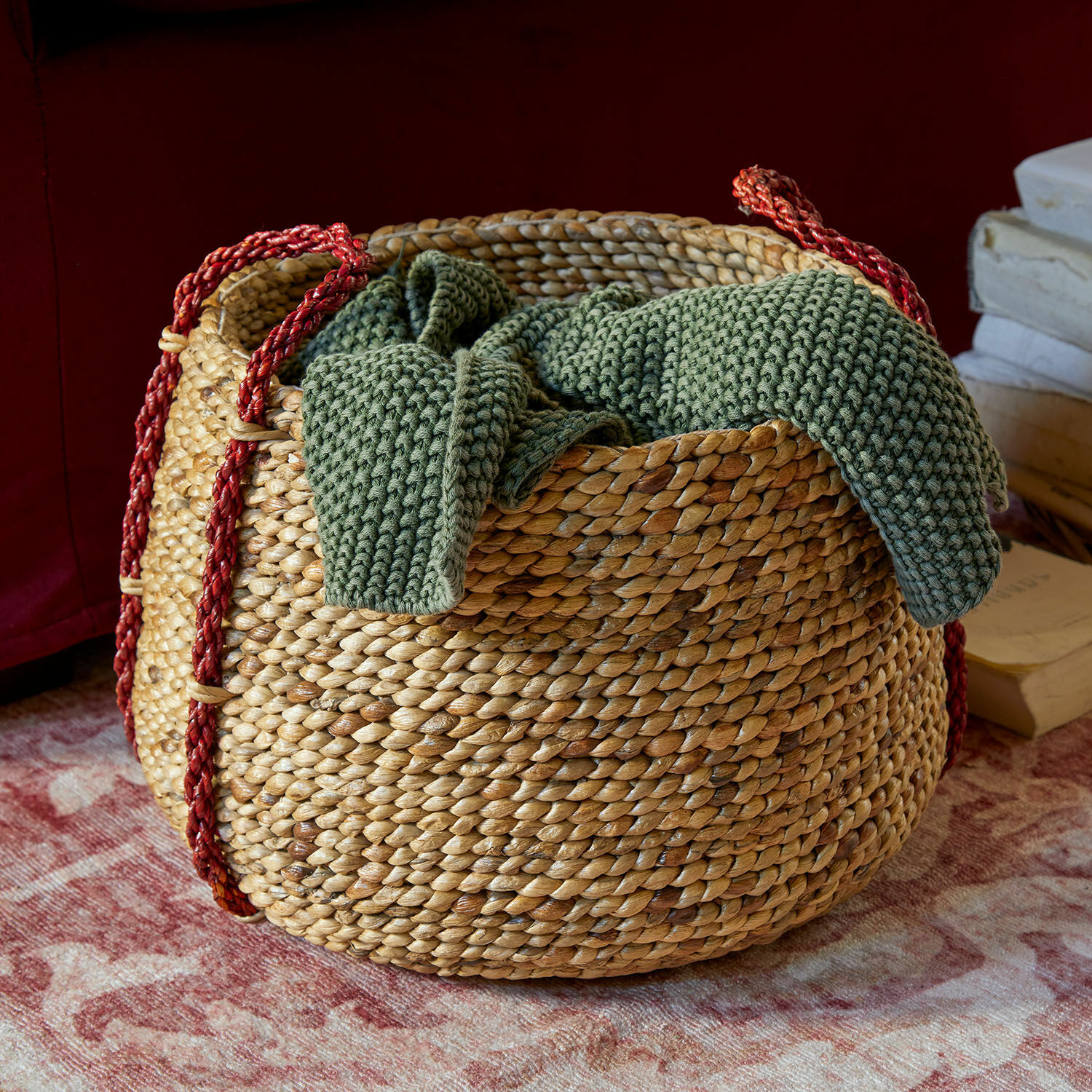 Cesta Cilovar Cesta redonda de mimbre con asas rojas y manta verde de punto sobre alfombra decorativa