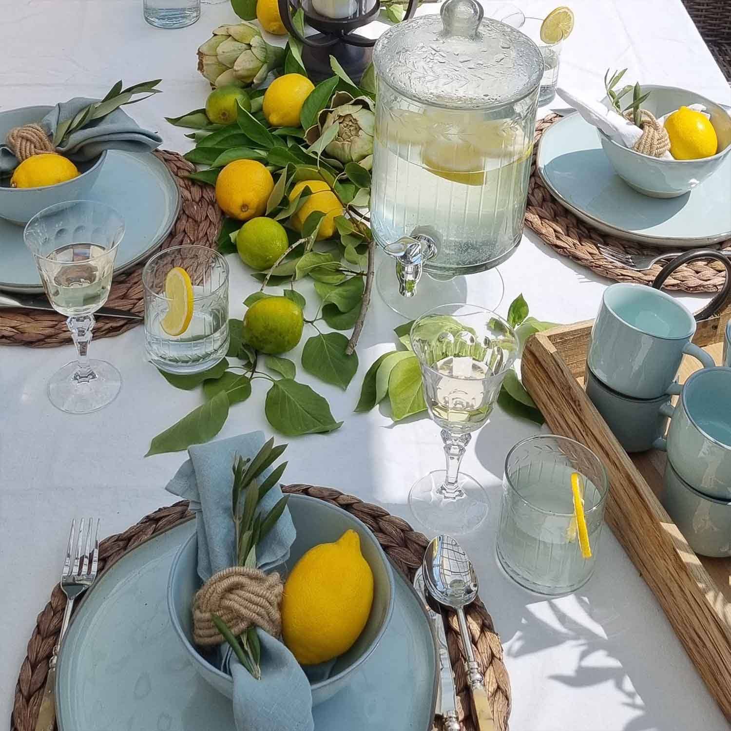 Mesa de verano con cerámica azul, limones y hojas verdes sobre mantel blanco, decoración de mesa