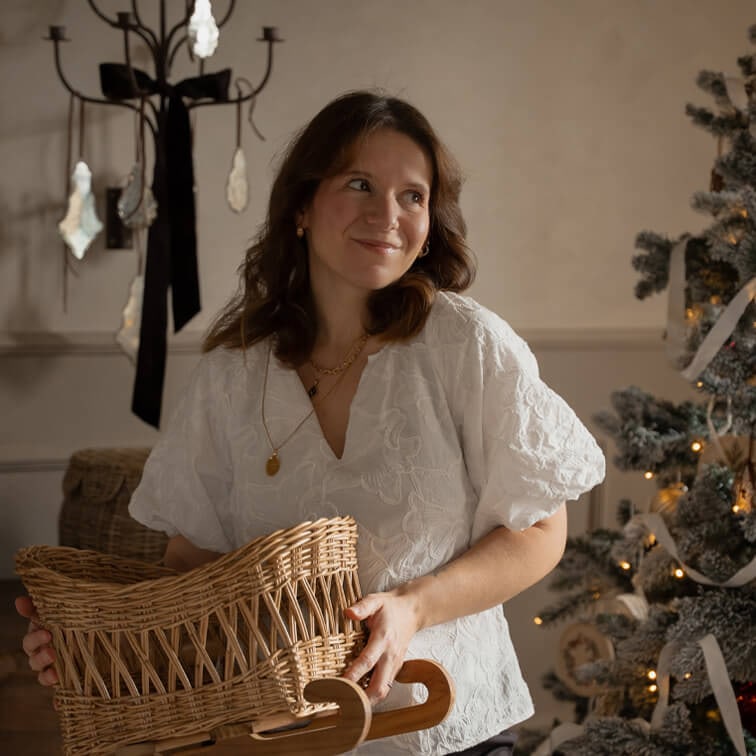 Mujer con blusa blanca bordada sosteniendo cesta de mimbre sobre trineo de madera junto a árbol de Navidad decorado