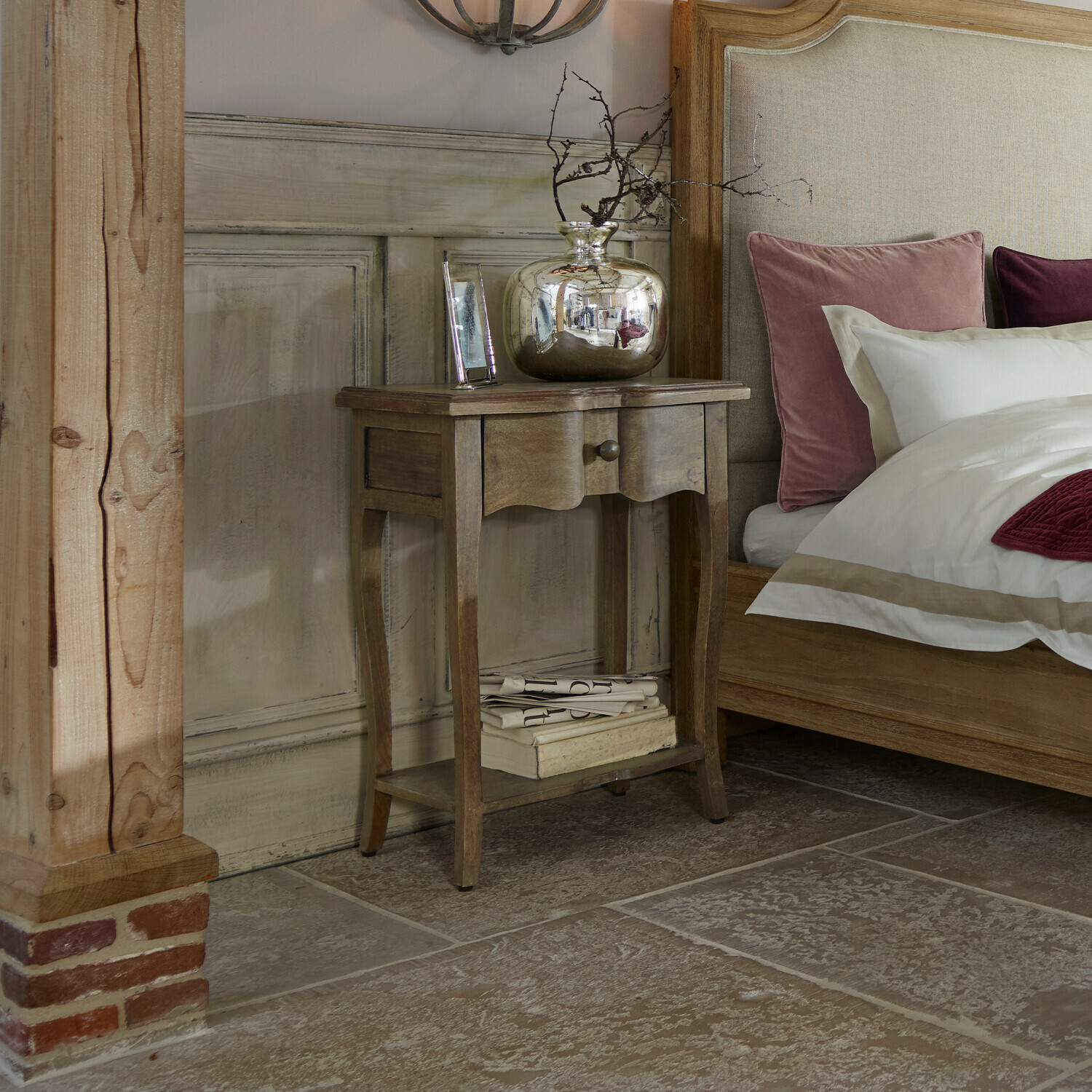 Mesa auxiliar Finstock Mesita de noche de madera con cajón y estante, decorada con jarrón plateado y libros junto a cama con cojines rosas