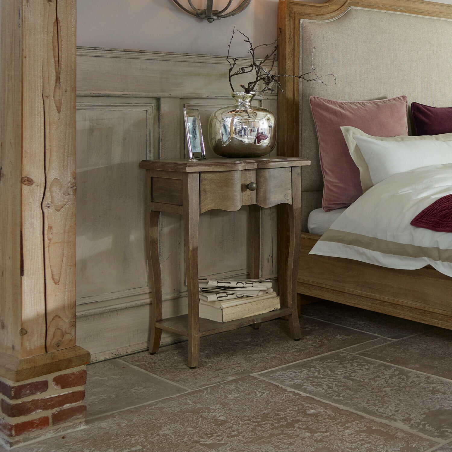 Mesa auxiliar Finstock Mesita de noche de madera con cajón y estante, decorada con jarrón plateado y libros junto a cama con cojines rosas