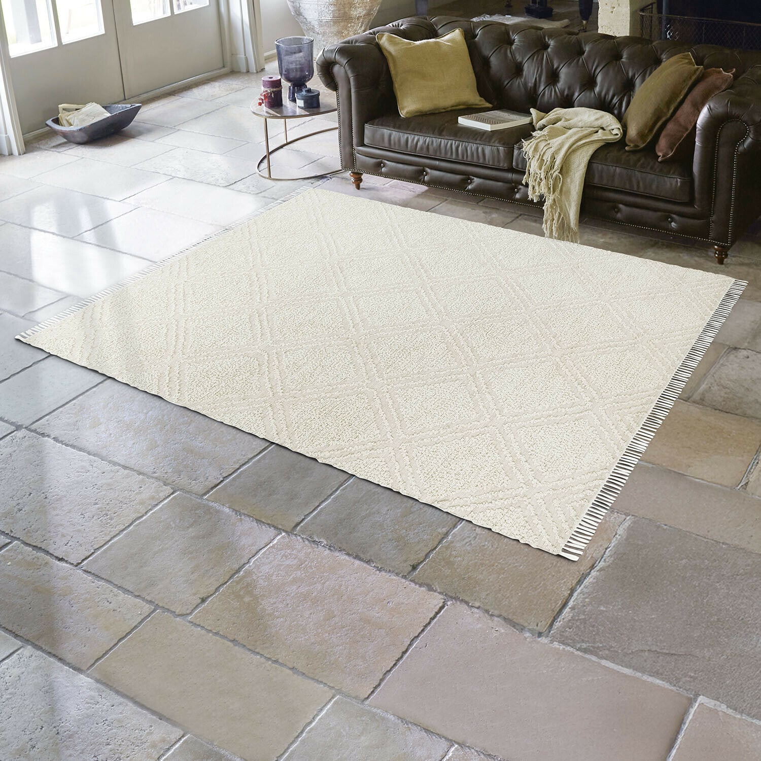 Alfombra para interior y exterior Bruand Alfombra beige con diseño geométrico sobre suelo de piedra en salón con sofá de cuero marrón