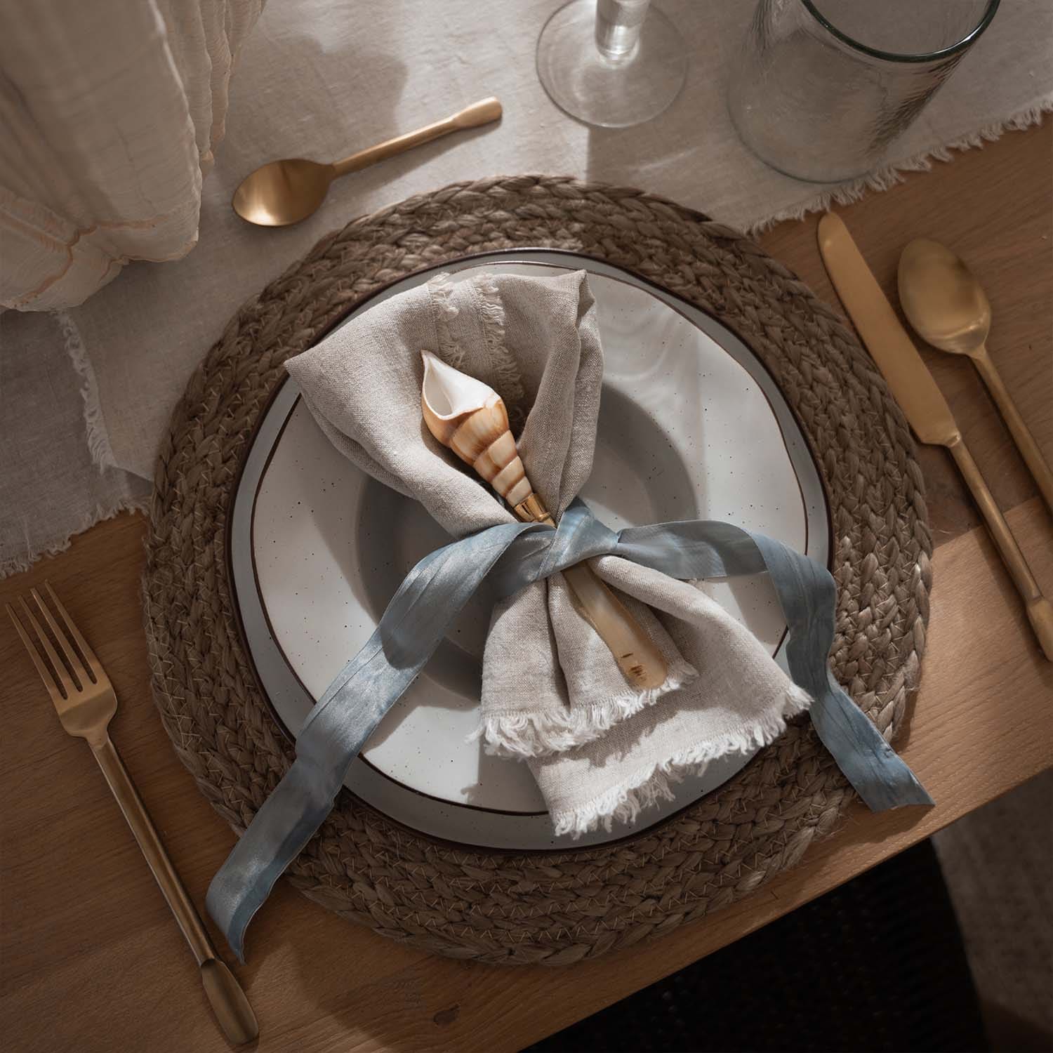 Mantel de lino natural con concha y cinta azul sobre plato con base trenzada y cubiertos dorados