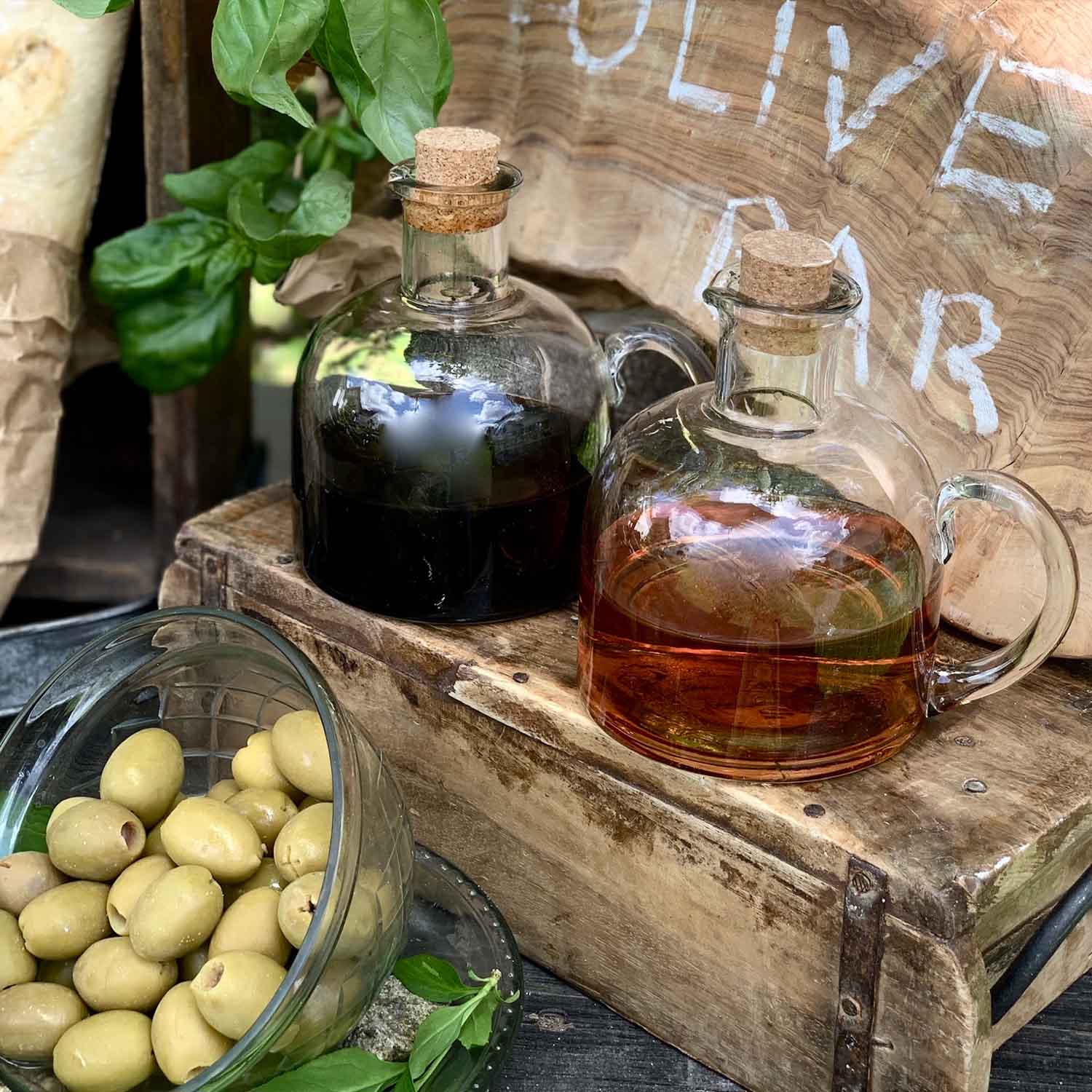 Botellas de vidrio con tapones de corcho con aceite y vinagre sobre caja de madera junto a aceitunas verdes y hojas de albahaca
