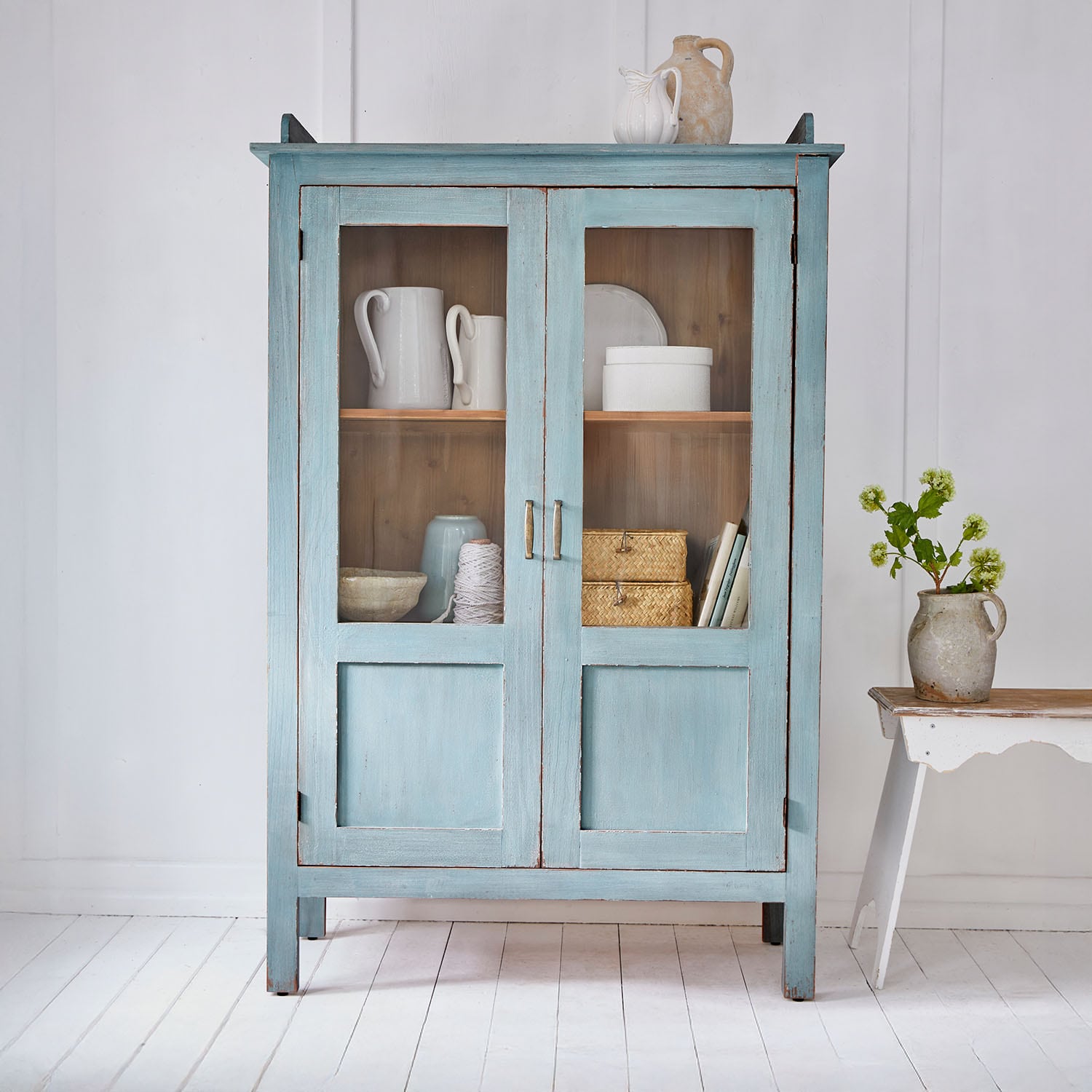 Armario de madera azul con puertas de cristal y decoraciones de cerámica vintage en habitación luminosa