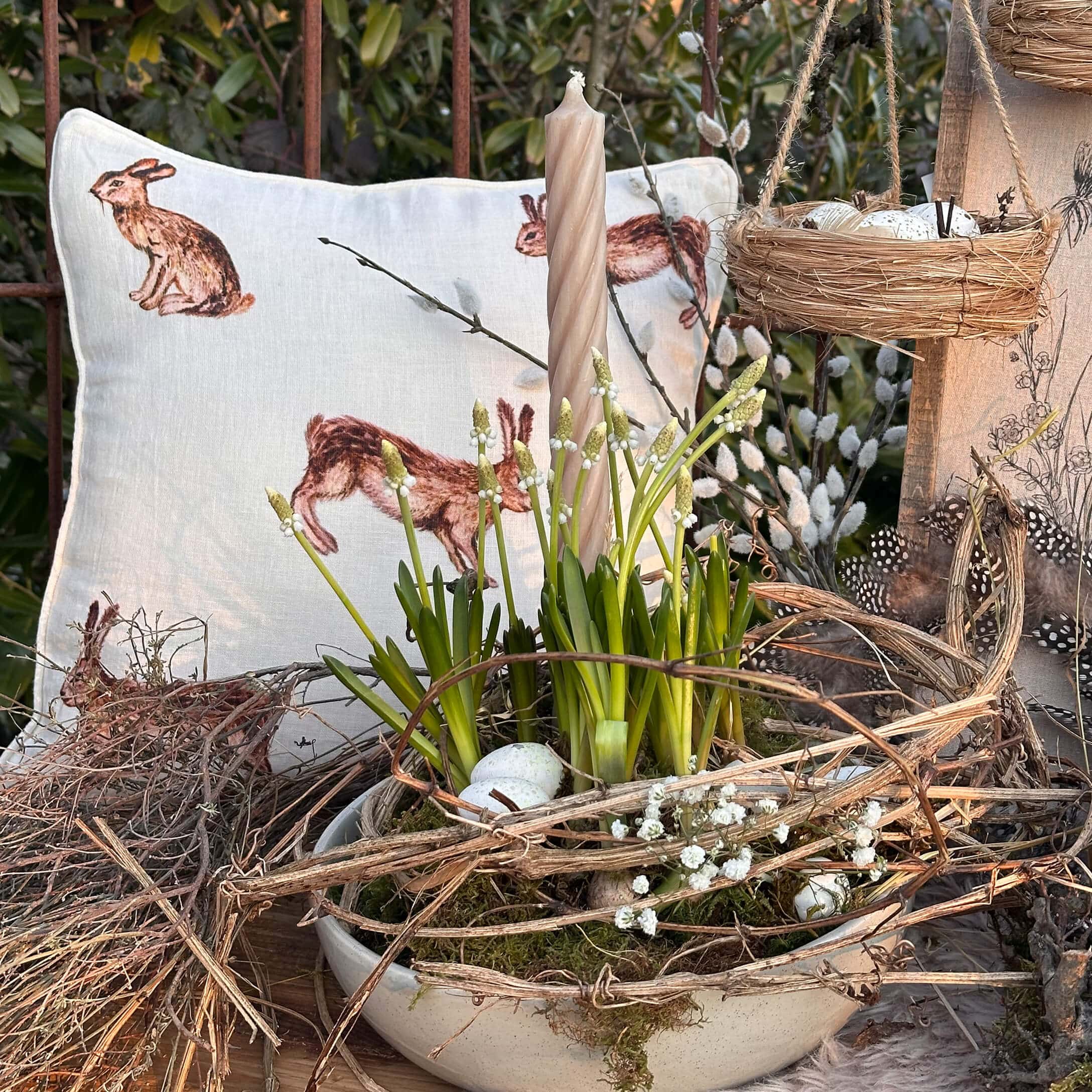Decoración de Pascua con vela blanca, flores verdes y cojín con diseño de conejo sobre mesa de madera