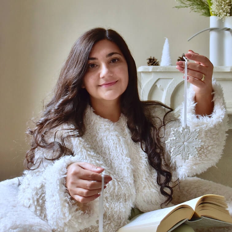 Mujer con suéter blanco y esponjoso sosteniendo adorno navideño blanco en forma de copo de nieve con libro abierto en las piernas