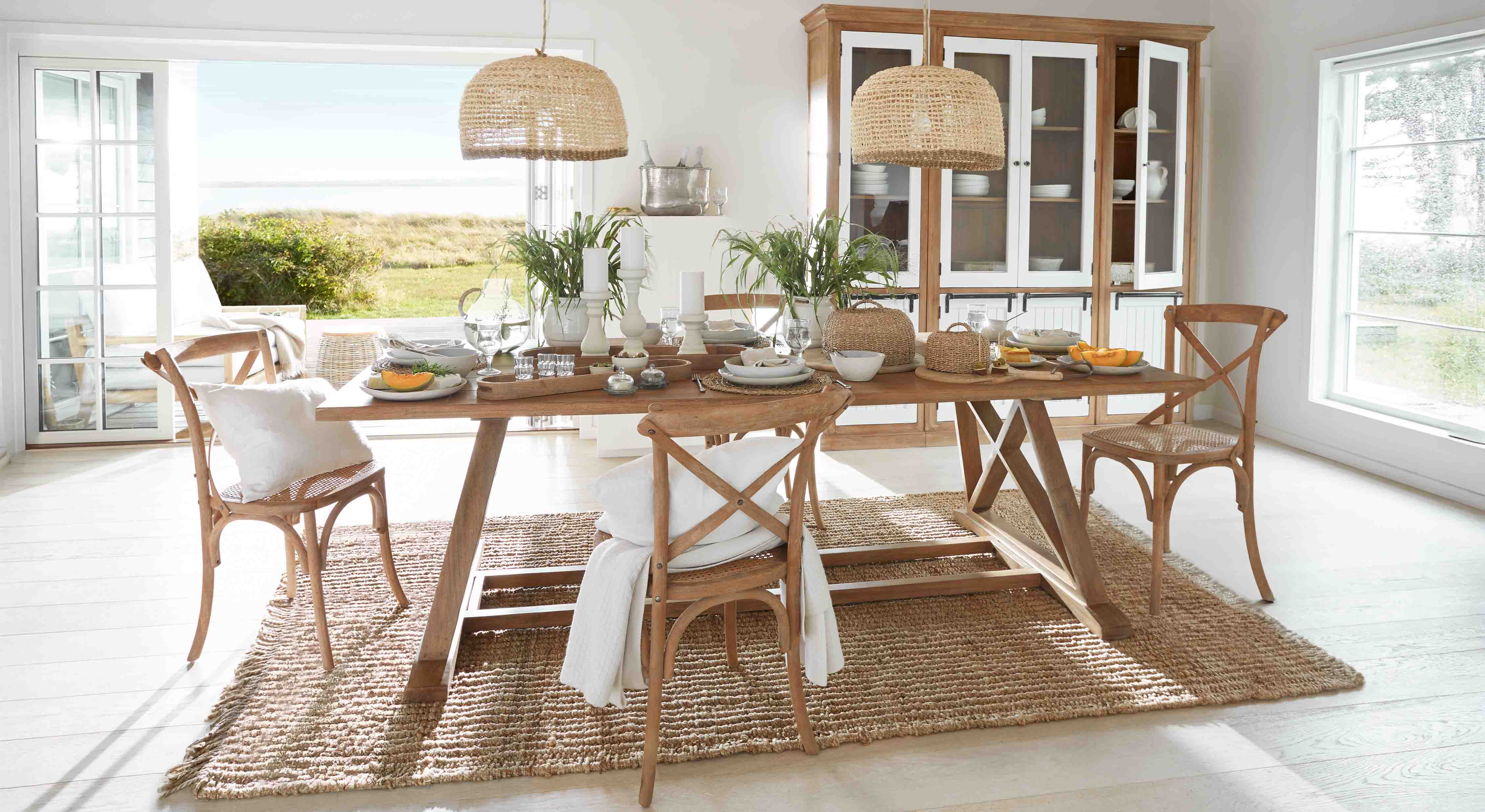 Comedor con mesa de madera y sillas de ratán sobre alfombra natural, decoración vegetal y lámparas de mimbre