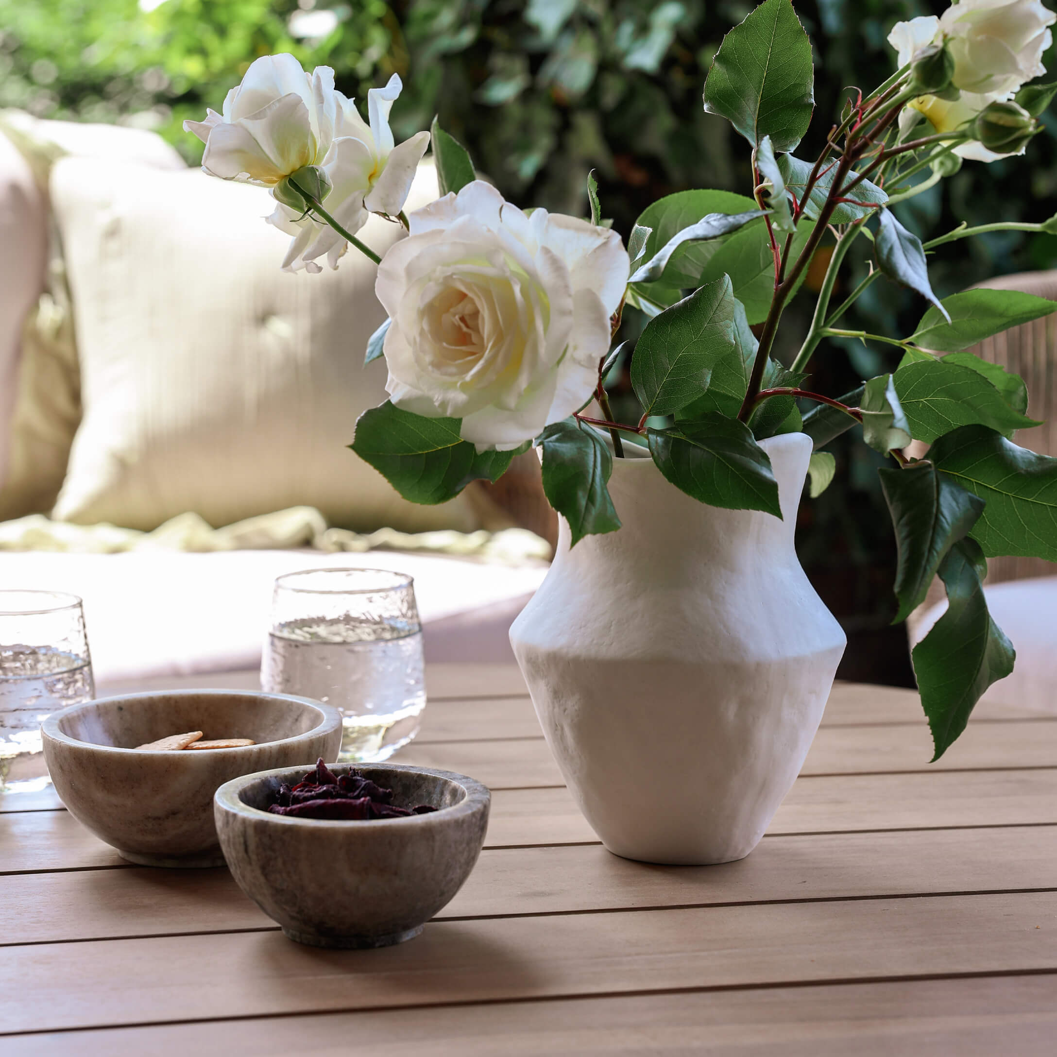 Jarrón de cerámica blanca con rosas blancas sobre mesa de madera con cuencos y vasos de agua