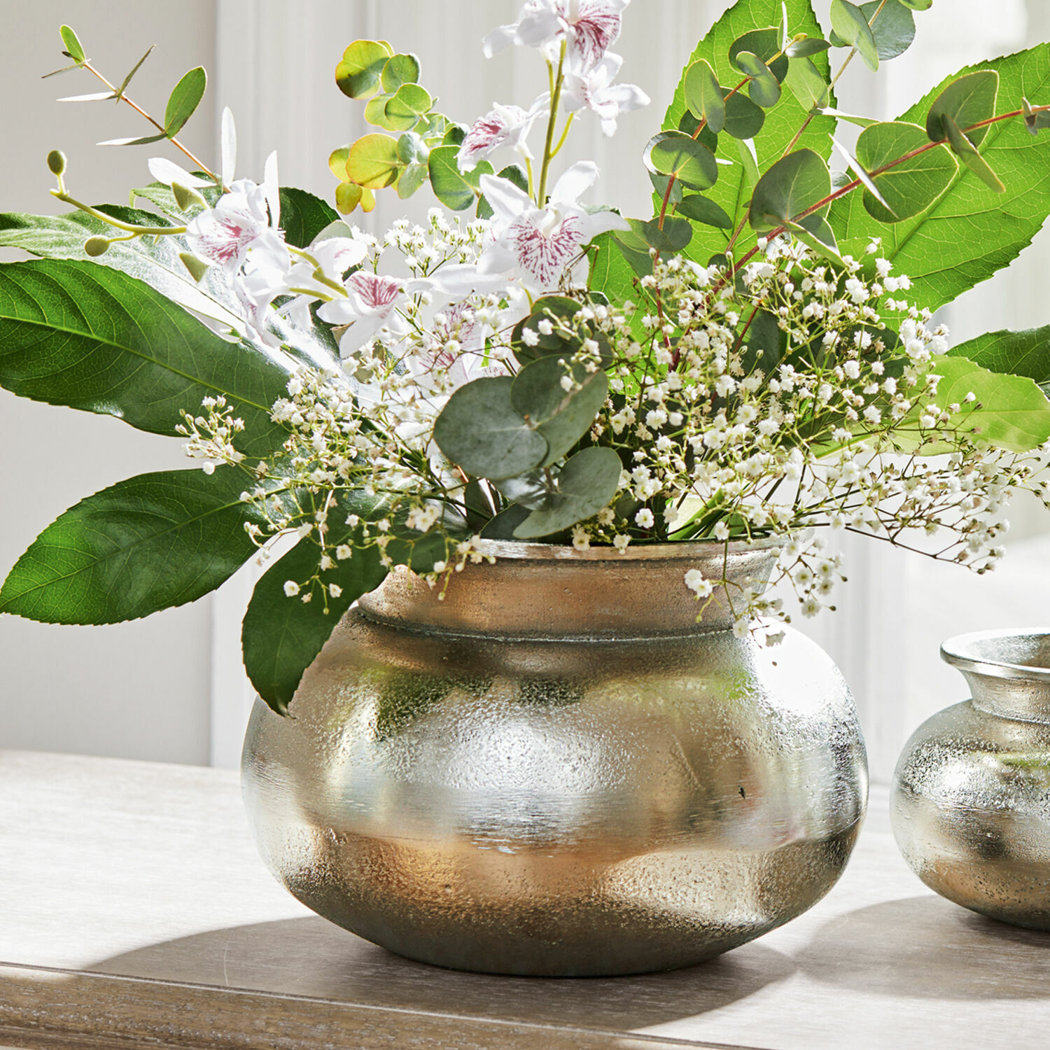 Jarrón Hadrien Jarrón metálico plateado con flores blancas y hojas verdes sobre mesa de madera, decoración interior
