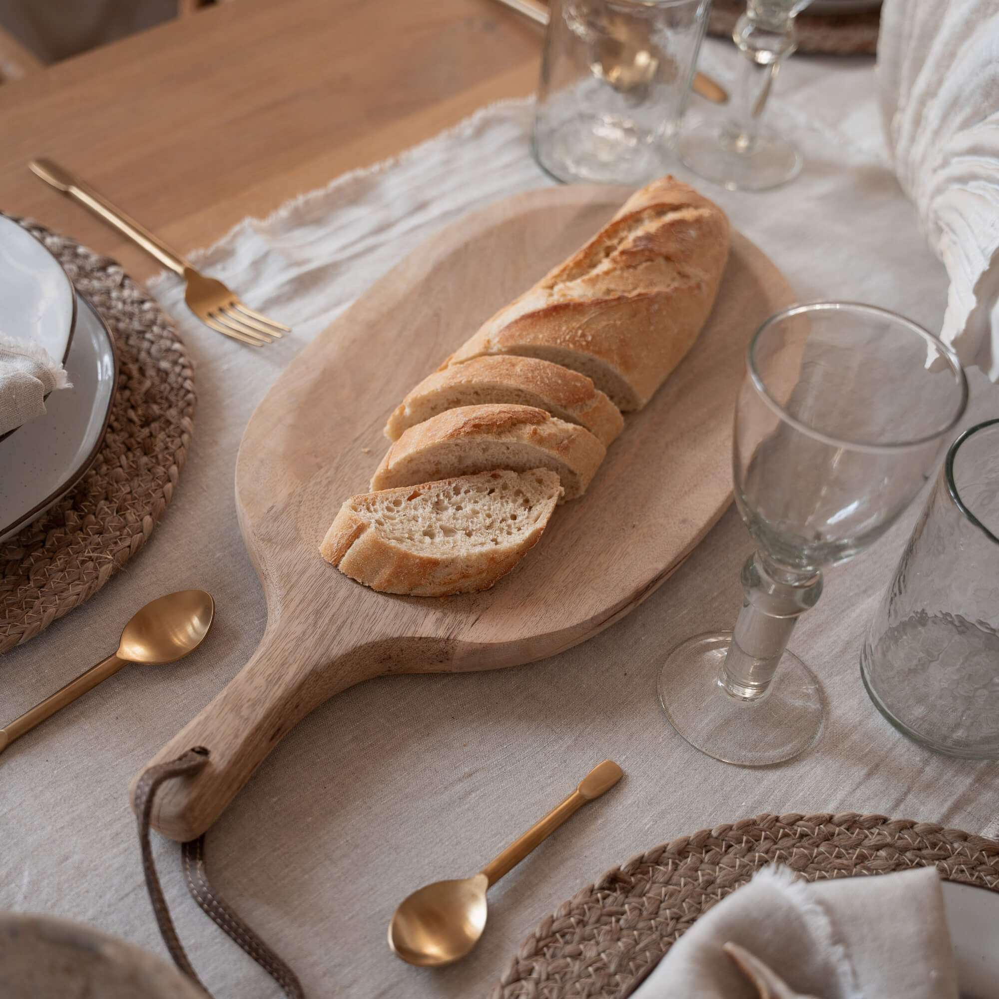 Tabla de cortar de madera con baguette cortada sobre mesa con vajilla y cubiertos dorados