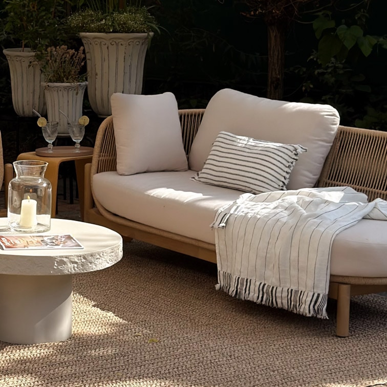 Sofá de exterior moderno con cojines claros y manta a rayas sobre alfombra natural en jardín