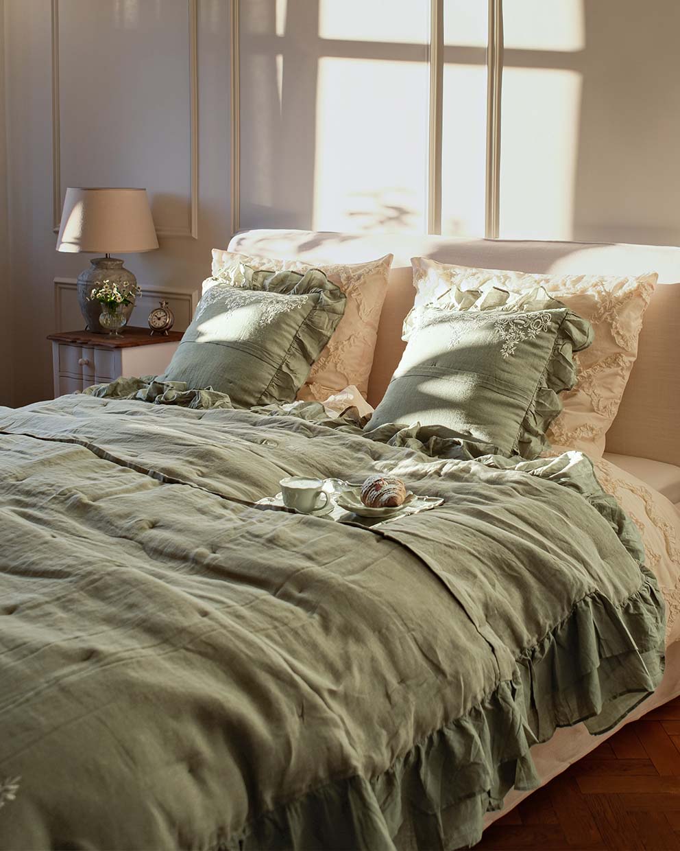 Ropa de cama verde de lino con volantes y cojines bordados en dormitorio luminoso con lámpara y reloj