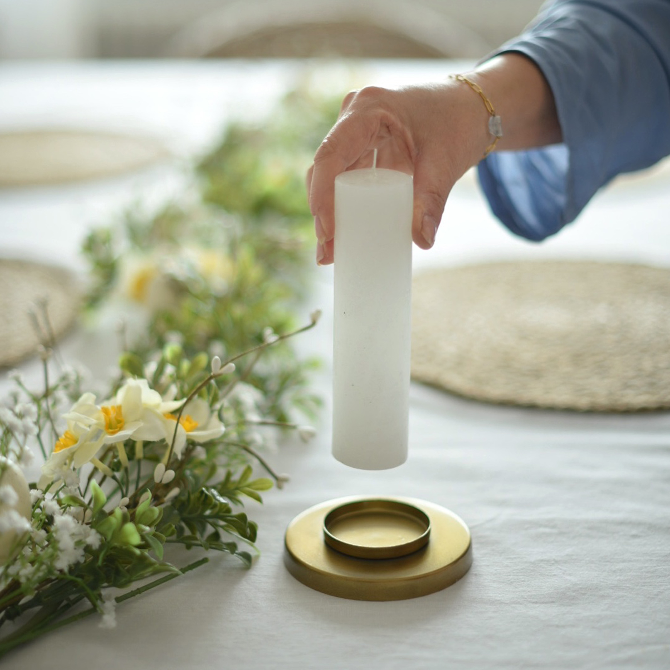 Vela blanca cilíndrica sostenida sobre un candelabro dorado con decoración floral sobre mantel blanco