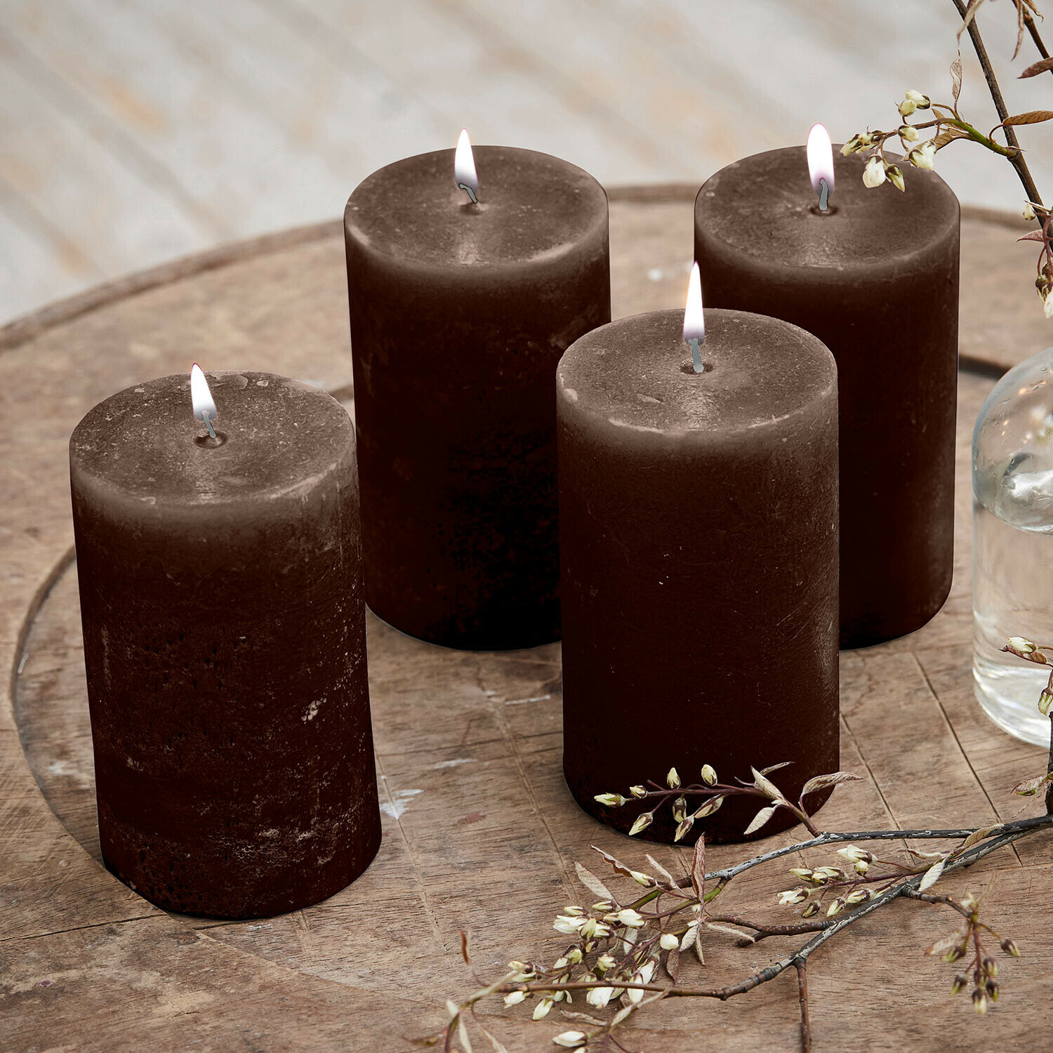 Set de 4 velas Braga Cuatro velas marrones de cera encendidas sobre una mesa de madera con ramas decorativas