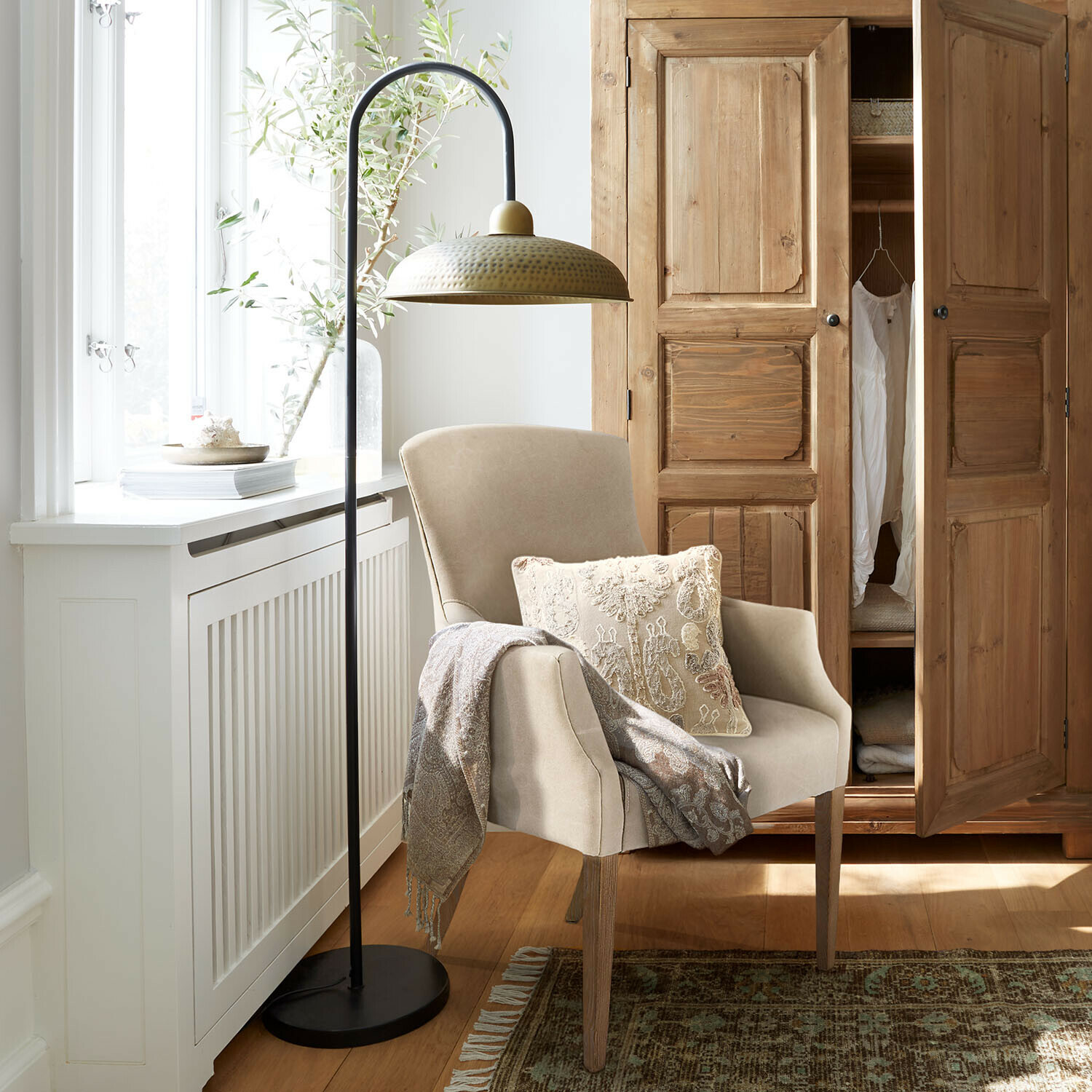 Sillón beige con patas de madera, cojín decorativo y manta, junto a lámpara de pie metálica con pantalla de latón