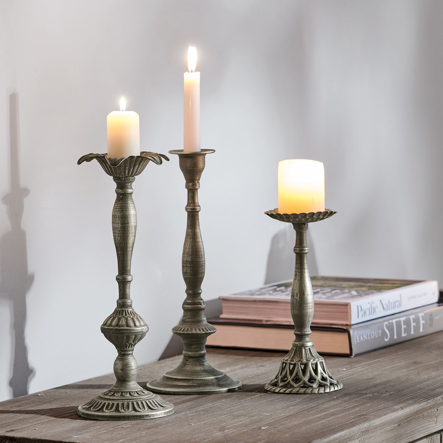 Set de 3 candeleros Chandra Tres candelabros de metal color plata envejecida con velas encendidas sobre mesa de madera