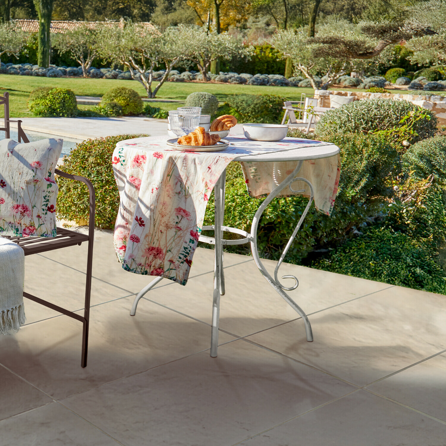 Mesa Aube Mesa de jardín redonda de metal blanco con mantel floral y bollería en terraza con vista al jardín