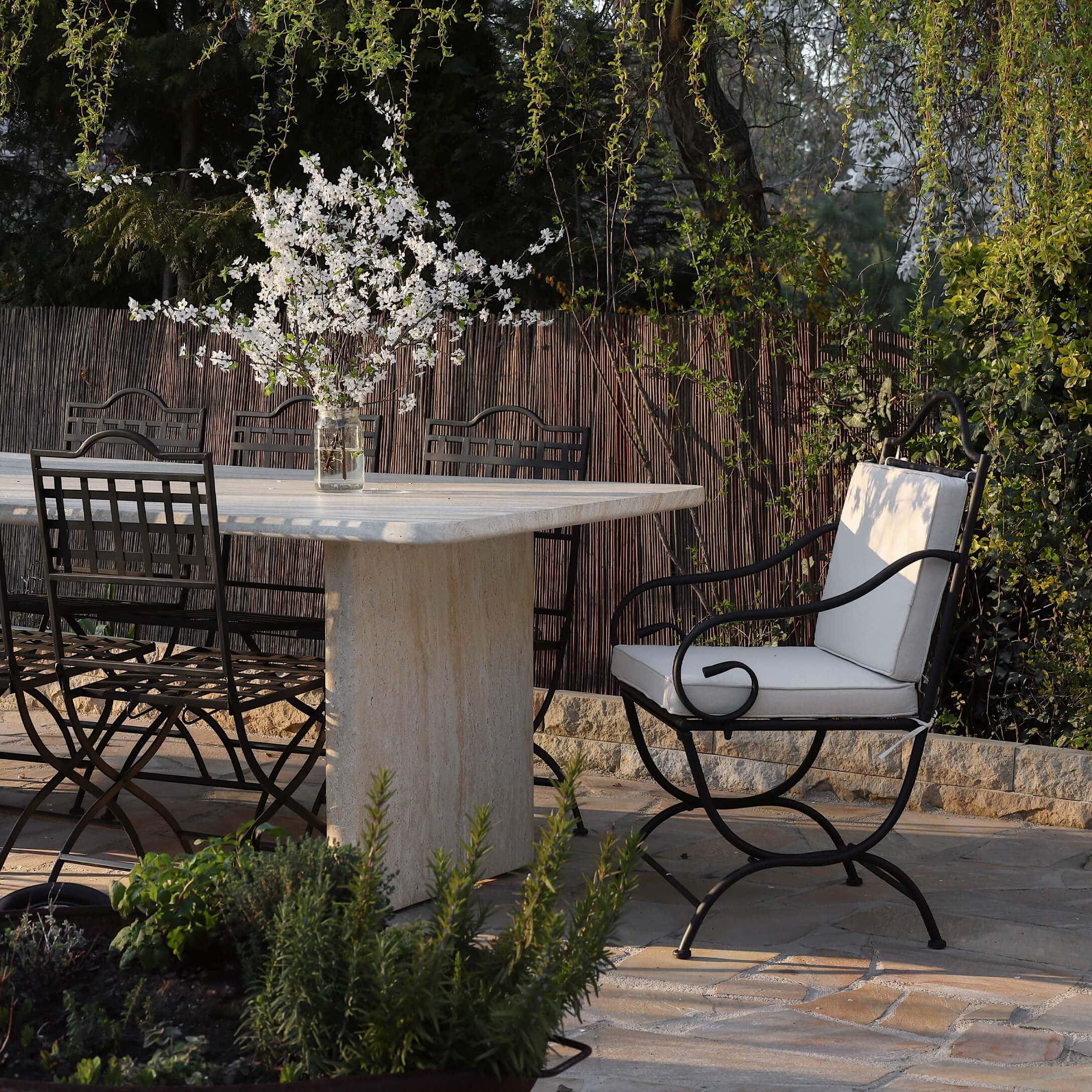 Mesa de piedra con sillas de metal negro y cojines claros en jardín con flores blancas