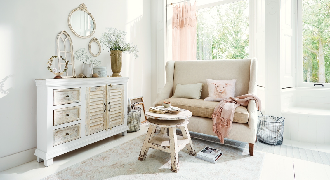 Salón acogedor estilo shabby chic con aparador de madera blanco y sofá beige con cojines y manta rosa