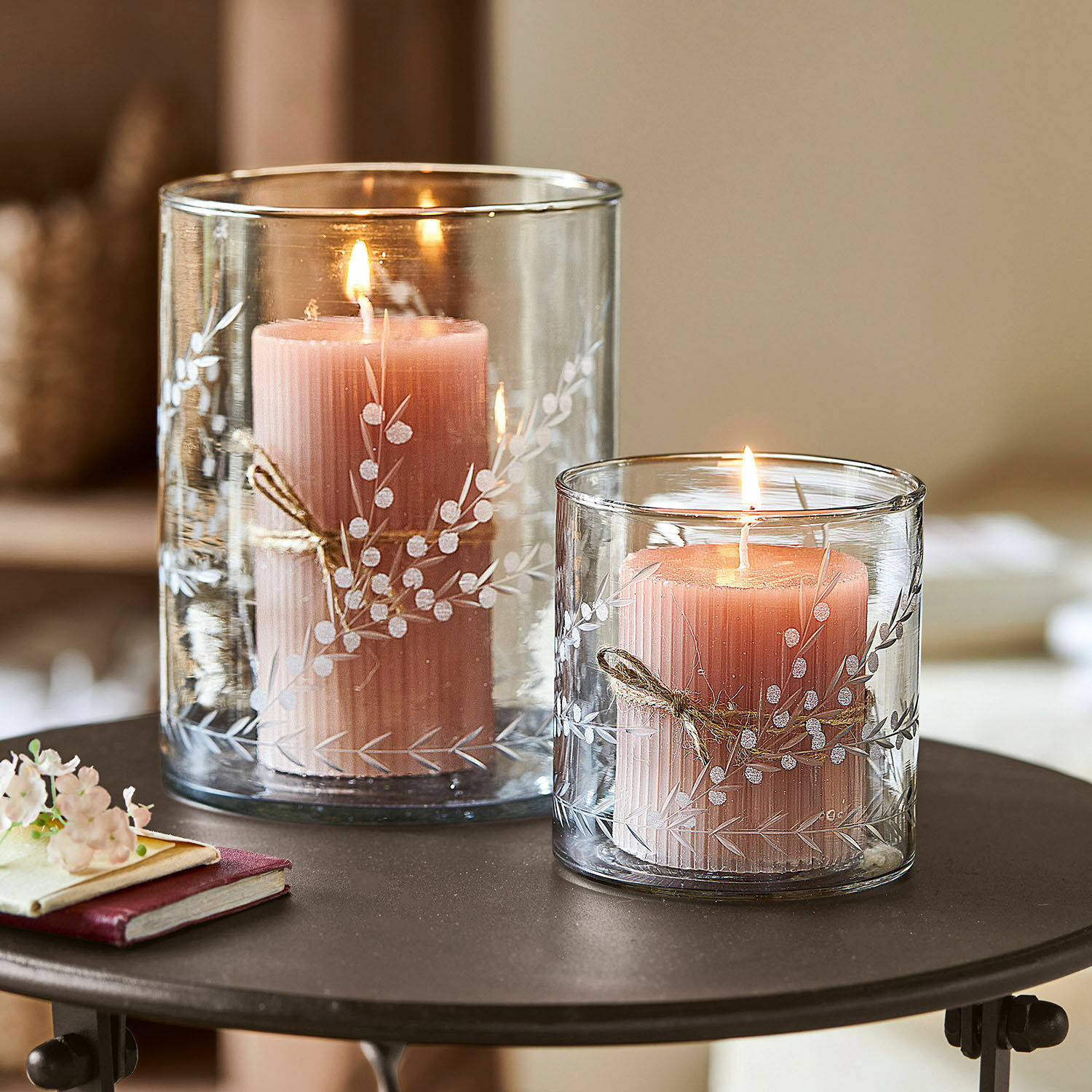 Dos velas rosas en portavelas de vidrio transparente con diseño vegetal blanco sobre una mesa
