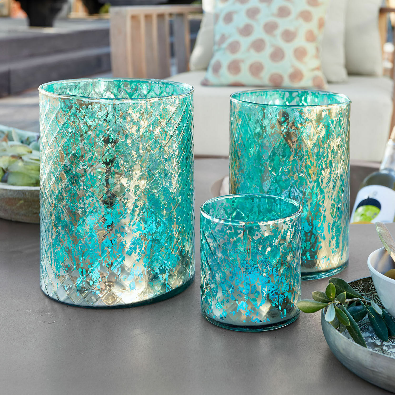 Set de 3 portavelas Firalen Conjunto de tres portavelas de vidrio turquesa con diseño metálico sobre mesa de jardín