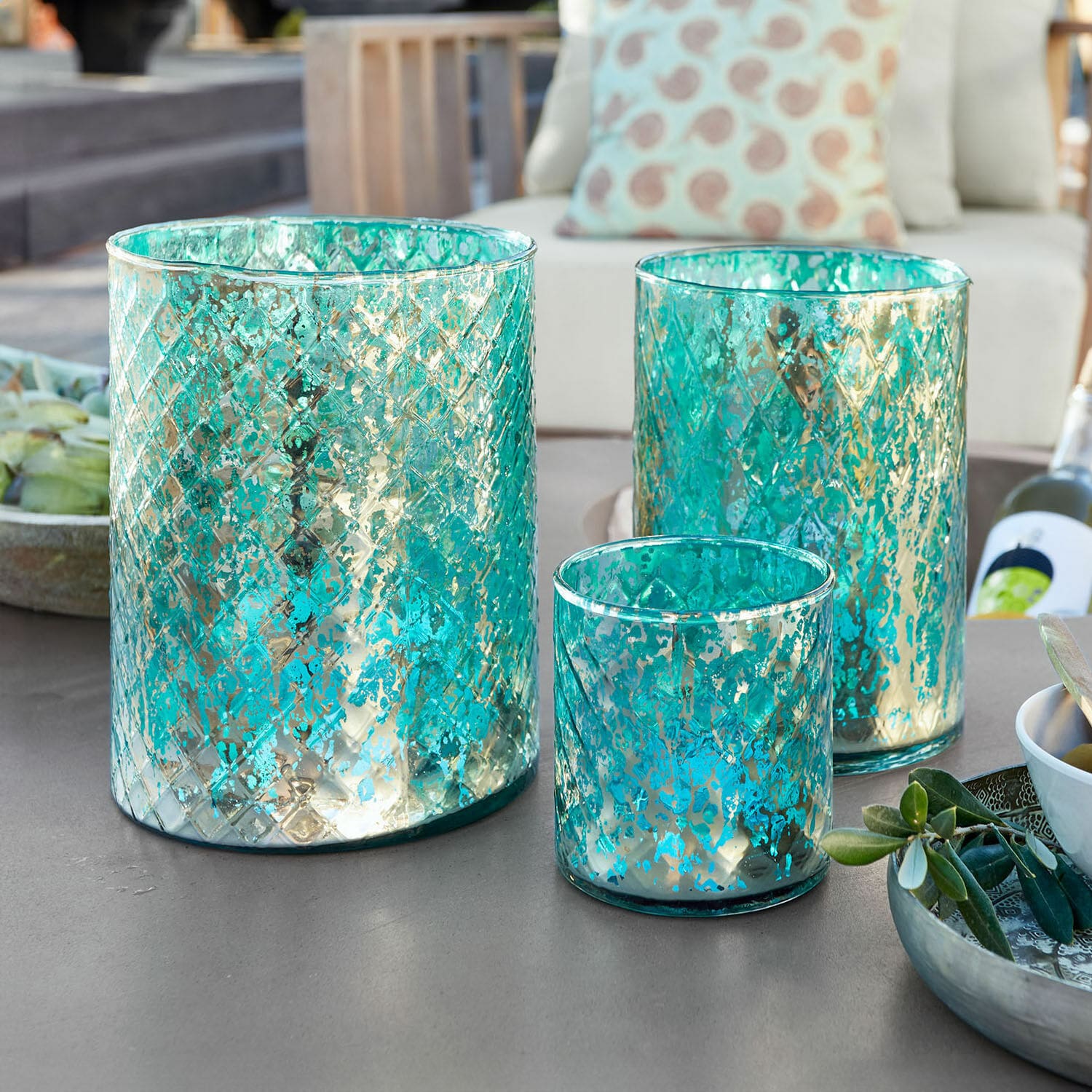 Set de 3 portavelas Firalen Conjunto de tres portavelas de vidrio turquesa con diseño metálico sobre mesa de jardín