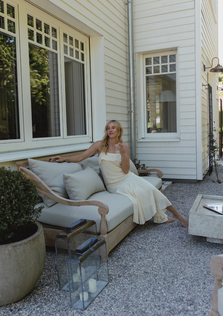 Sofá de jardín de madera con cojines gris claro en patio de grava con mujer con vestido crema