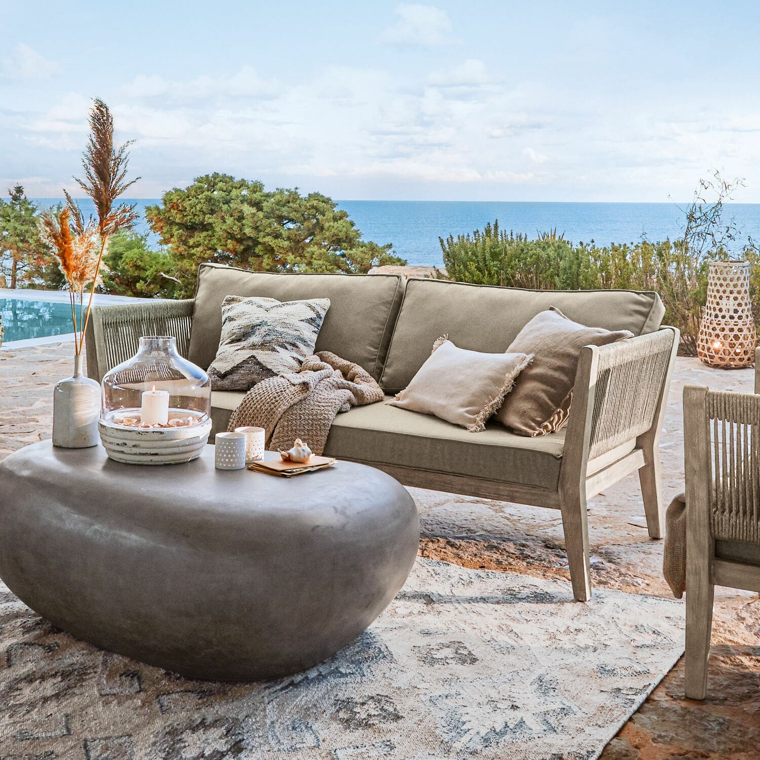 Sofá Biarritz Sofá de exterior beige de madera y tela en terraza con vista al mar, mesa de centro moderna