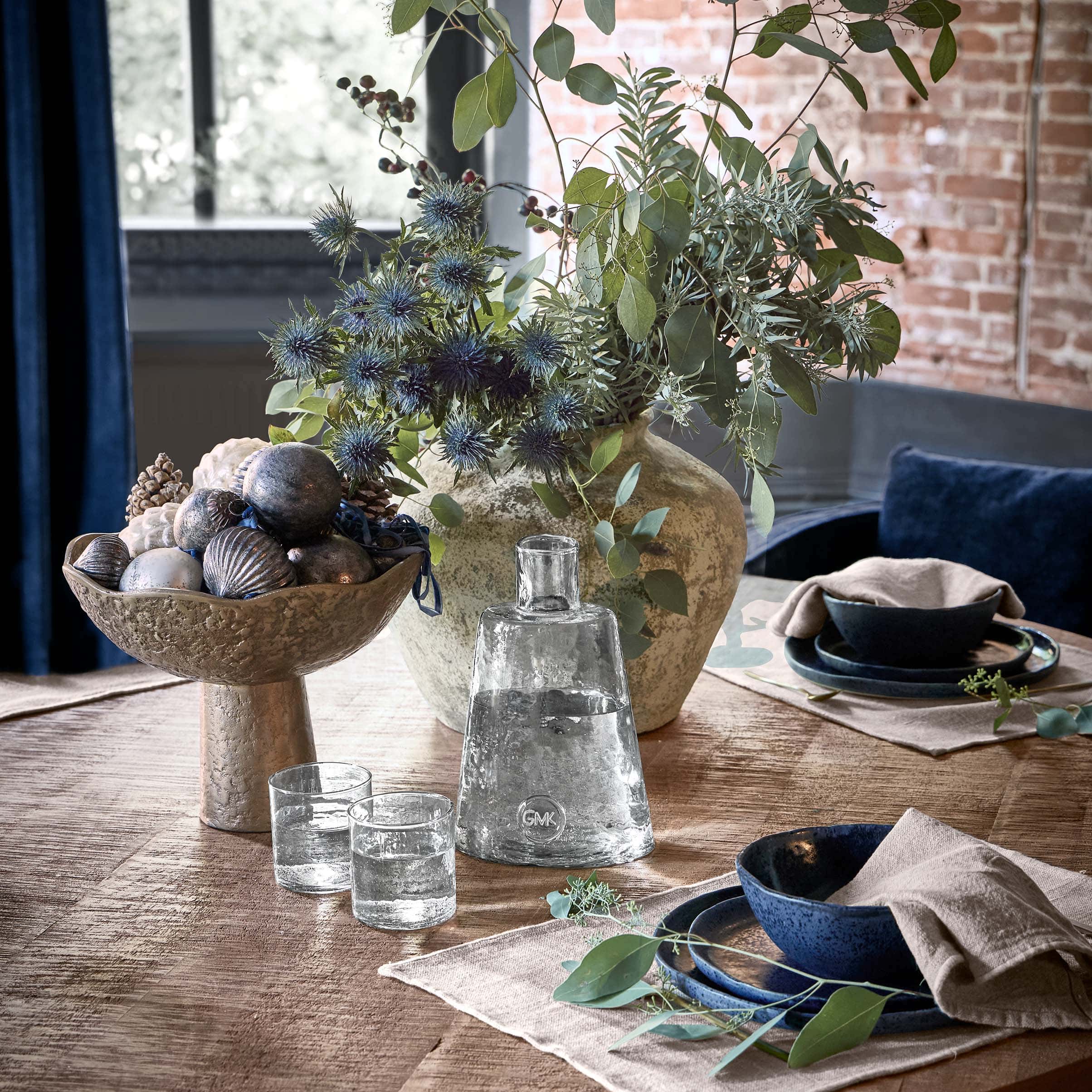 Cuenco decorativo de metal con adornos y jarrón con hojas verdes sobre mesa de madera en salón