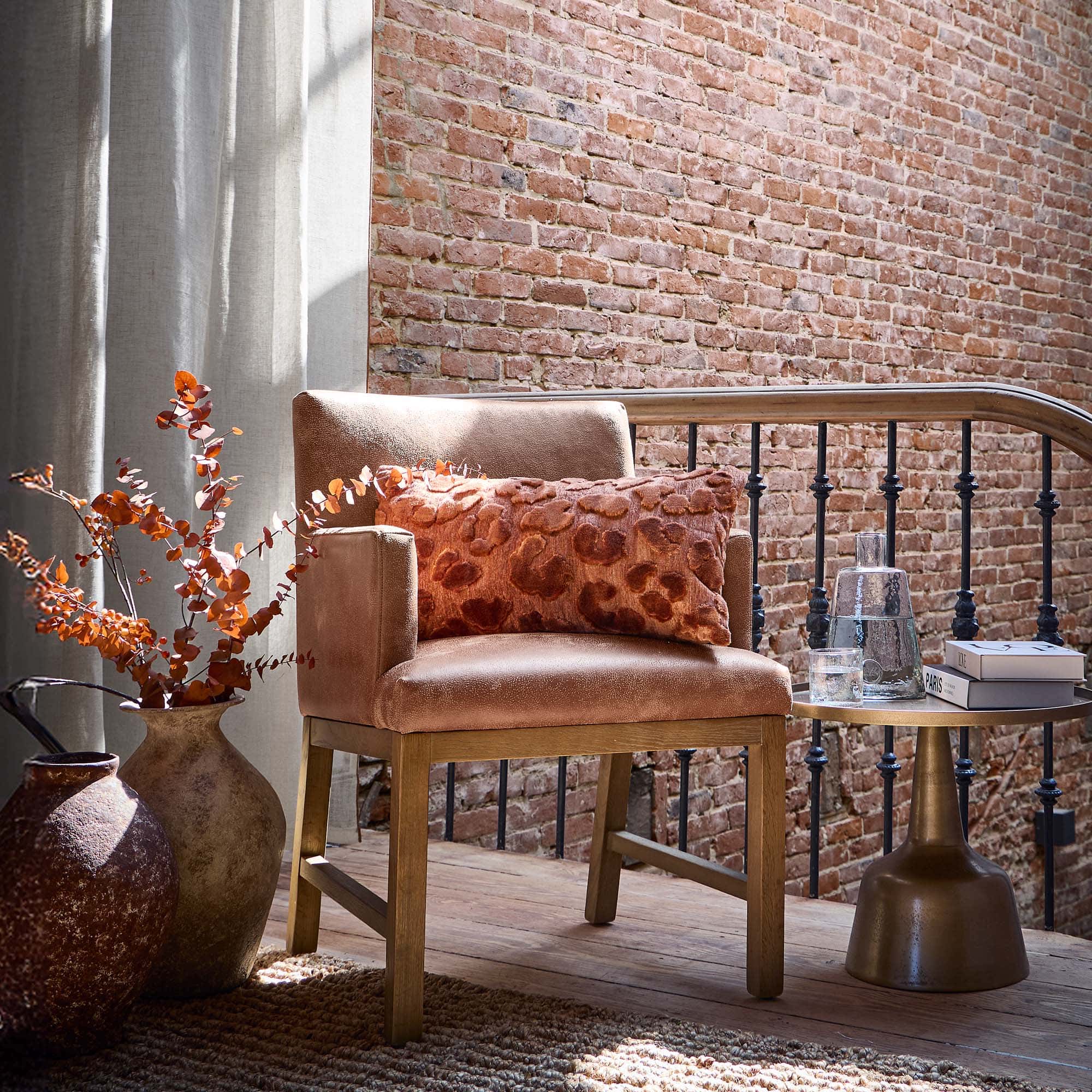 Sillón beige con base de madera y cojín naranja frente a pared de ladrillo y cortinas