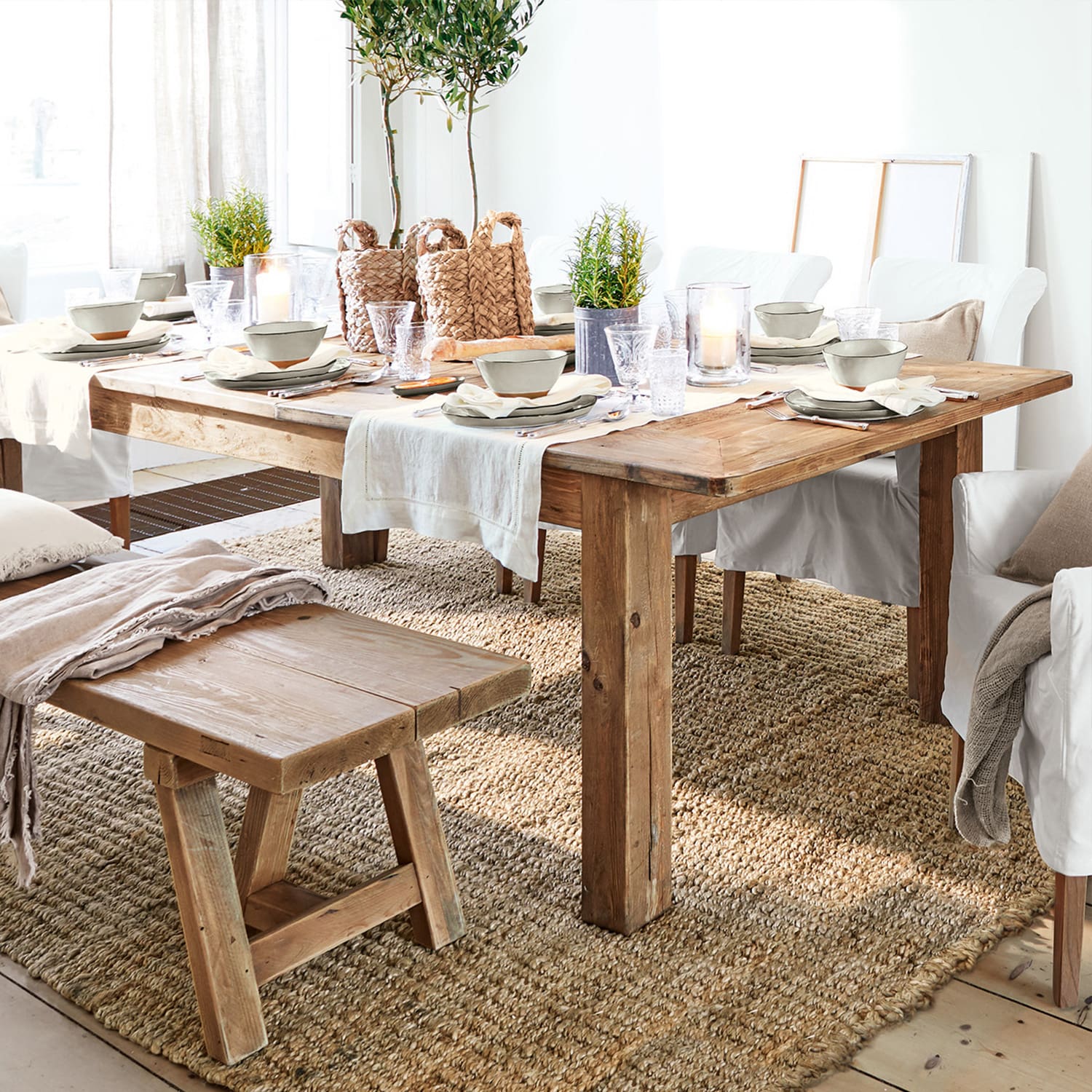 Mesa y banco rústicos de madera sobre alfombra natural con decoración de mesa y sillas blancas