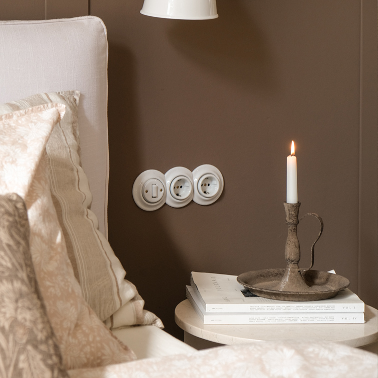 Candelero de metal rústico con vela blanca sobre mesa junto a cama con ropa de cama beige