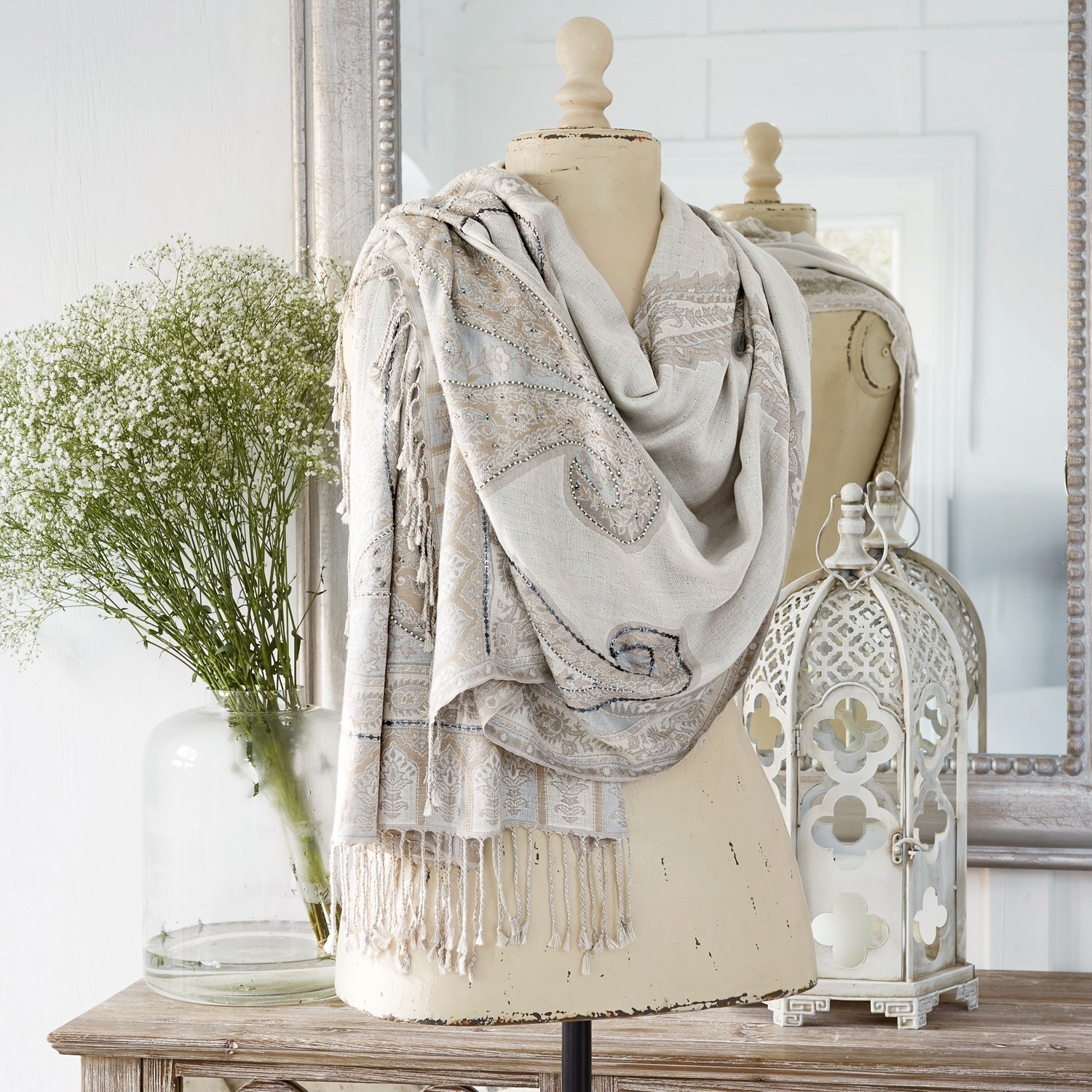 Chal Moyra Chal beige con diseño paisley y flecos en maniquí, decoración interior con farol y flores