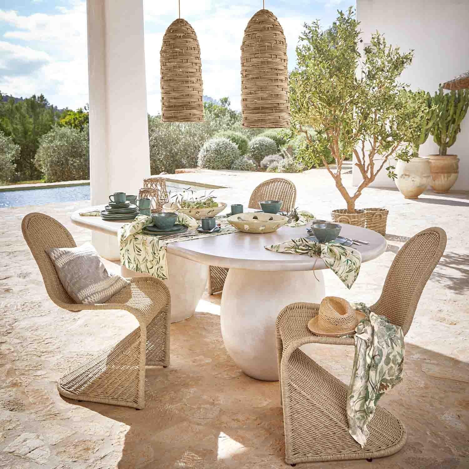 Mesa ovalada blanca de piedra con sillas de mimbre y lámparas colgantes naturales en jardín soleado