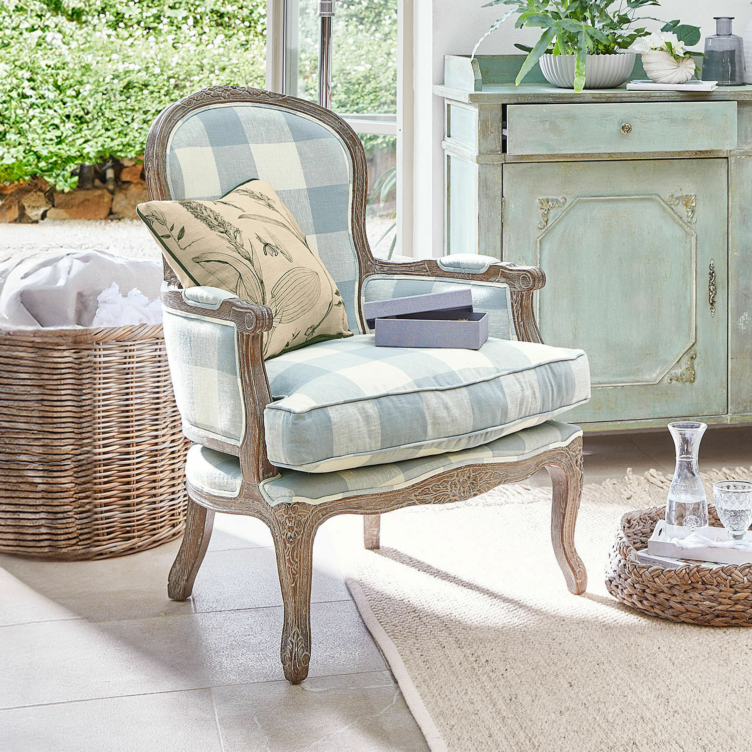 Sillón Quintelais Sillón de madera con tapizado a cuadros azul y beige, cojín decorativo con diseño vegetal en ambiente luminoso