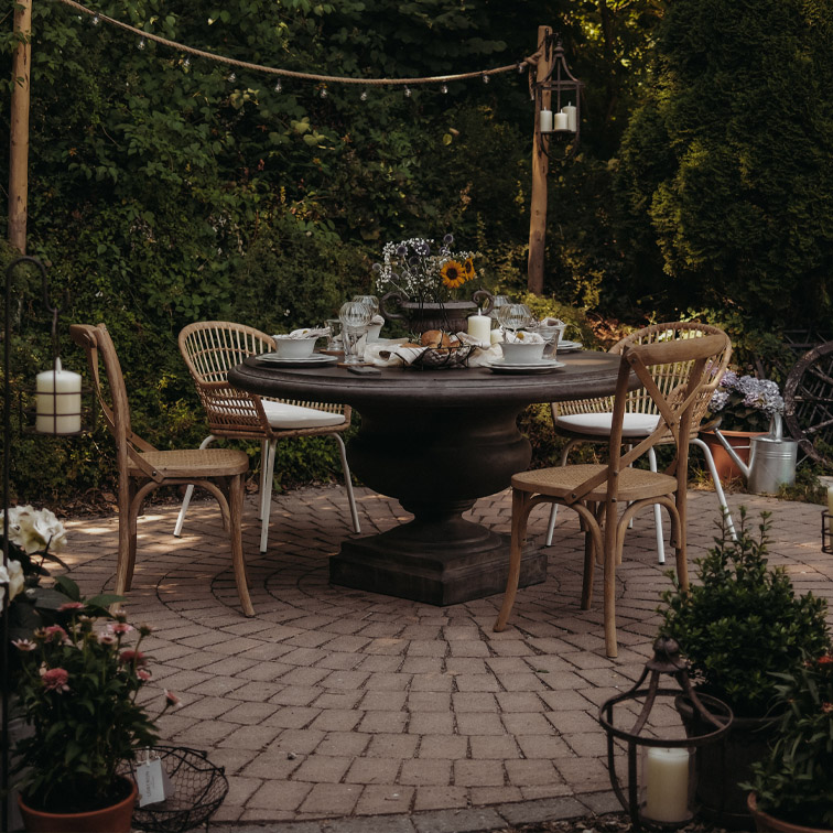 Mesa redonda de madera oscura con sillas de mimbre en patio adoquinado decorado con velas y flores al aire libre