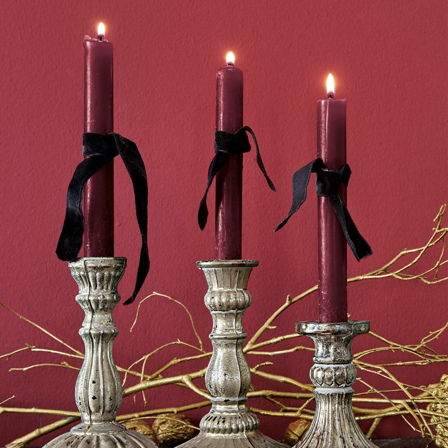Set de 12 velas Bauceau Tres candelabros metálicos plateados con velas burdeos y lazos negros sobre fondo rojo