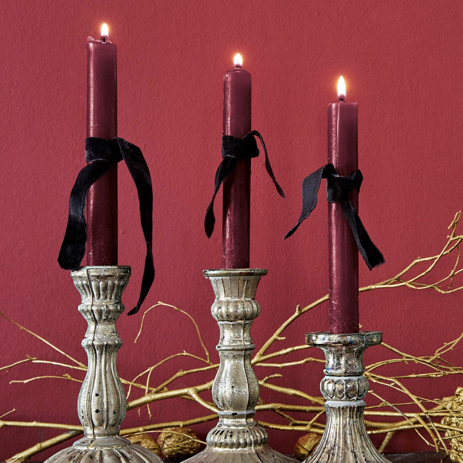 Set de 12 velas Bauceau Tres candelabros metálicos plateados con velas burdeos y lazos negros sobre fondo rojo