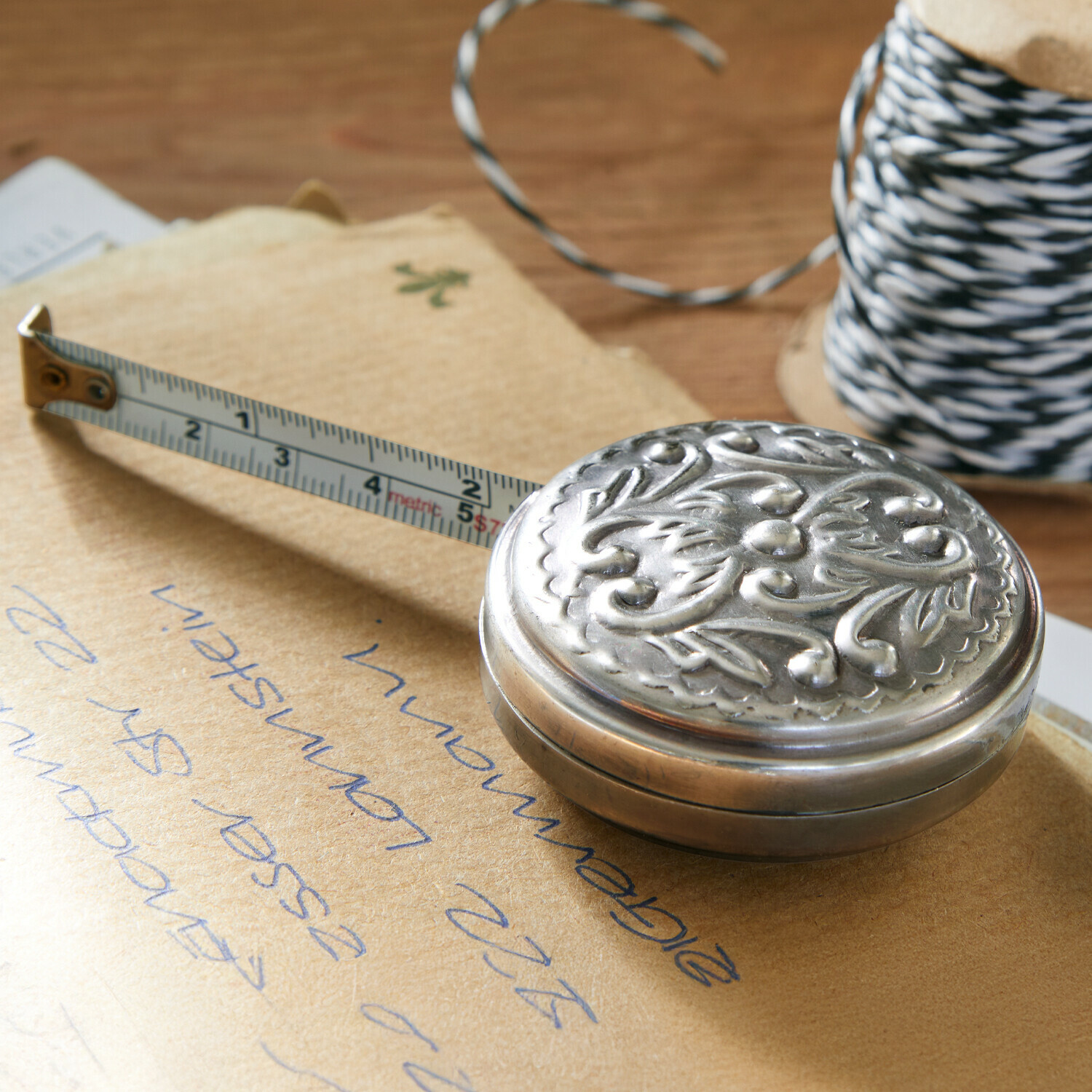 Cinta métrica Dakota Cinta métrica metálica decorativa con diseño vegetal sobre mesa de madera junto a carrete de cuerda