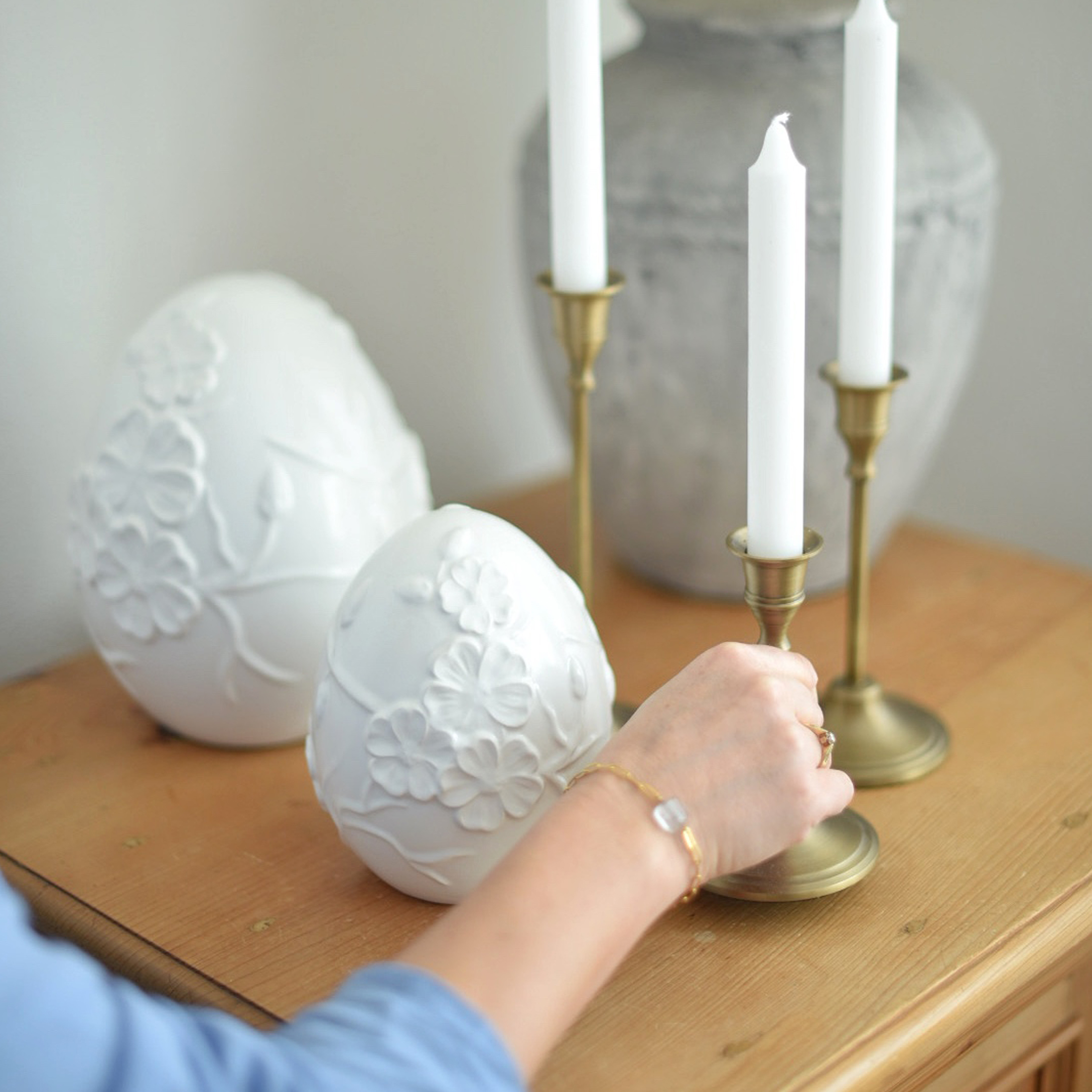 Huevos decorativos de cerámica blanca con relieve floral sobre mesa de madera junto a candelabros de latón