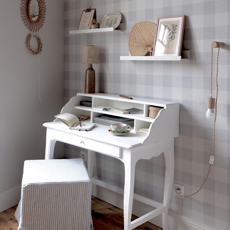 Escritorio blanco de madera con estantes, lámpara y taza en habitación acogedora con papel pintado a cuadros